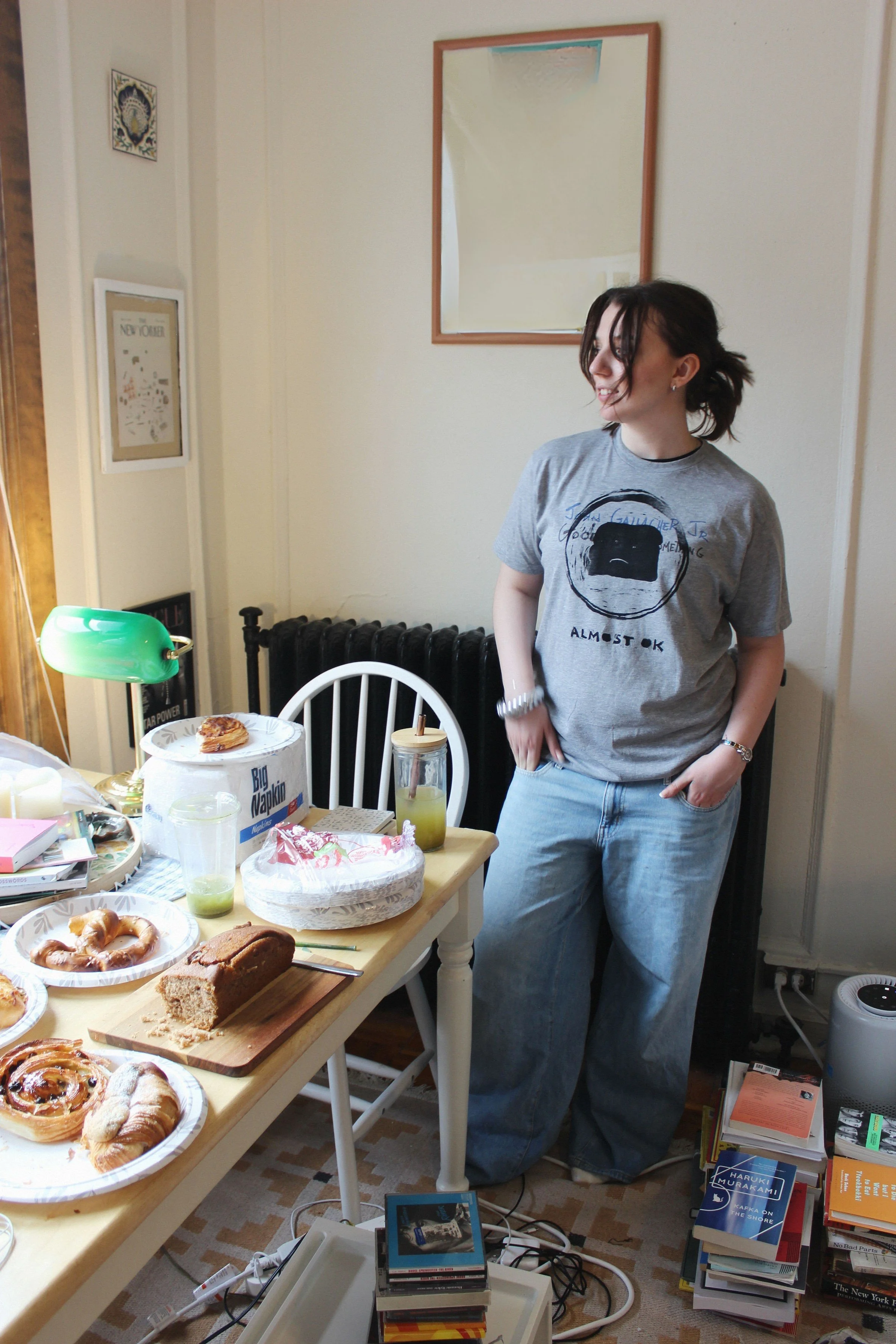 A woman standing in a room with a table filled with various baked goods and drinks. She is wearing a gray T-shirt and loose jeans, smiling and looking to her right. The room has a white wall with a mirror above a radiator, and various books and items