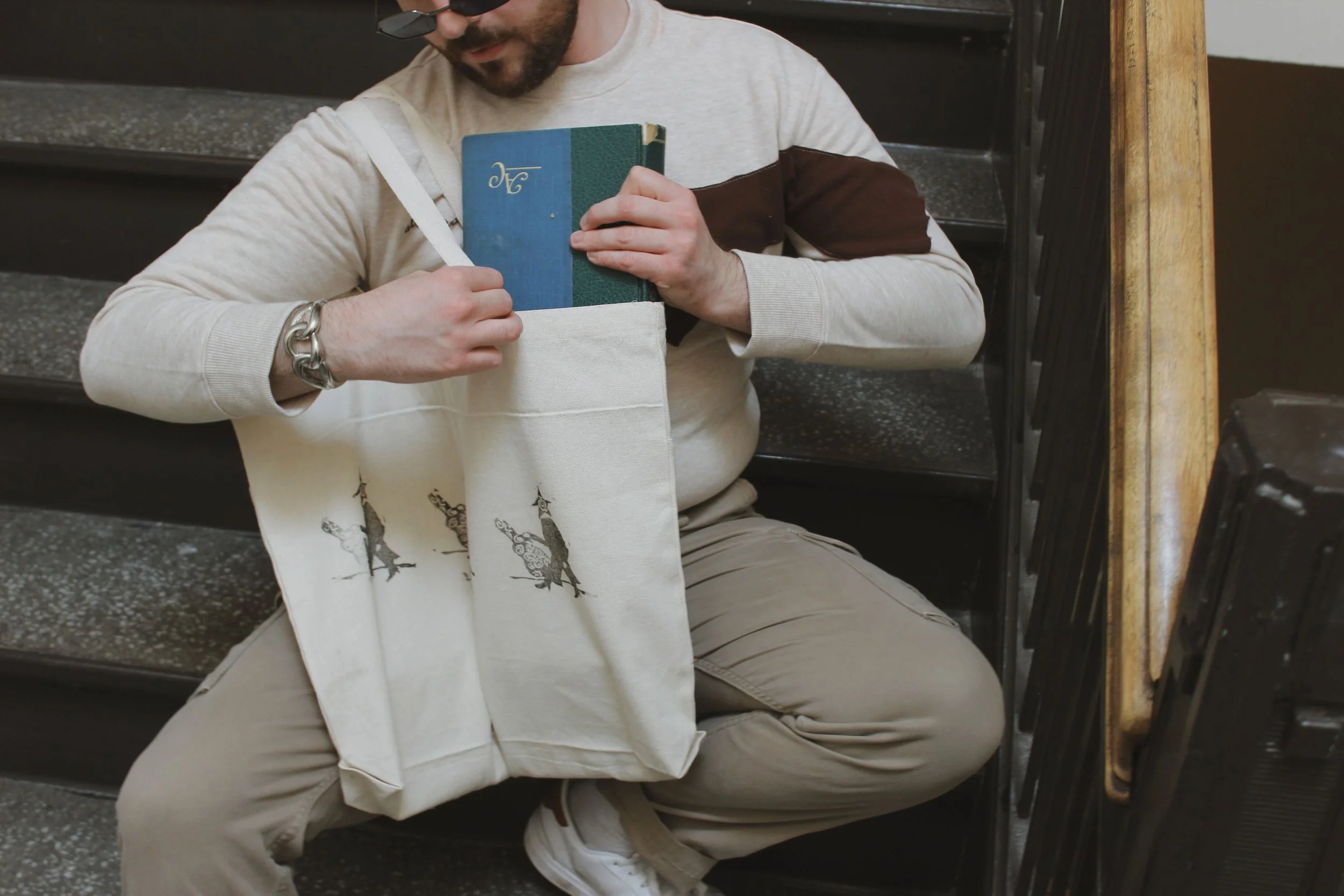 A man sitting on stairs puts a book into a white tote bag with animal illustrations, wearing beige pants, a light-colored sweatshirt with brown patches, a bracelet, and glasses.