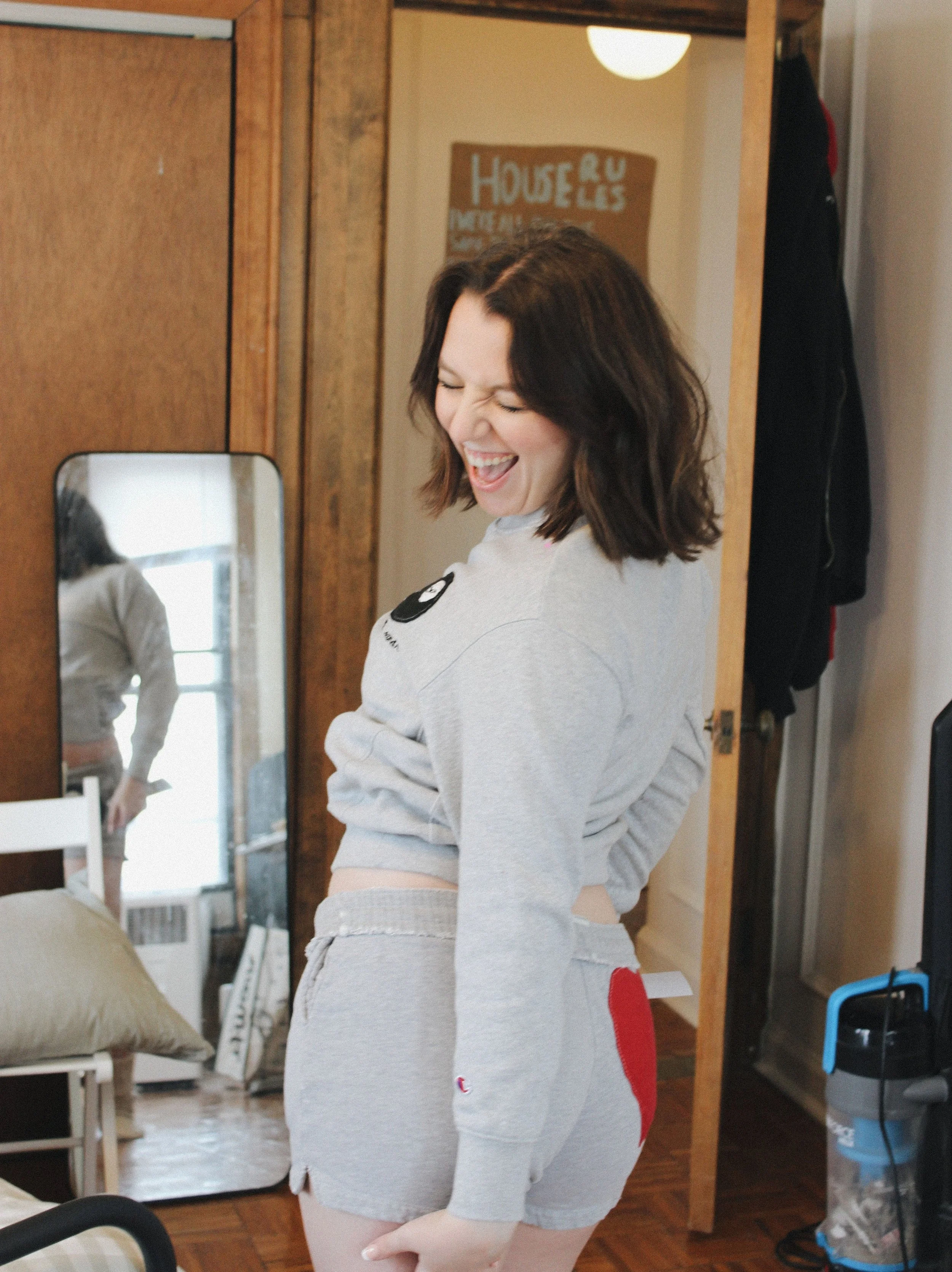 A woman with shoulder-length dark hair is smiling and laughing in a cozy, wood-paneled room. She's wearing a light gray sweatshirt and matching shorts. There is a mirror behind her showing her back, and furniture, a window, and a poster on the wall are also visible.