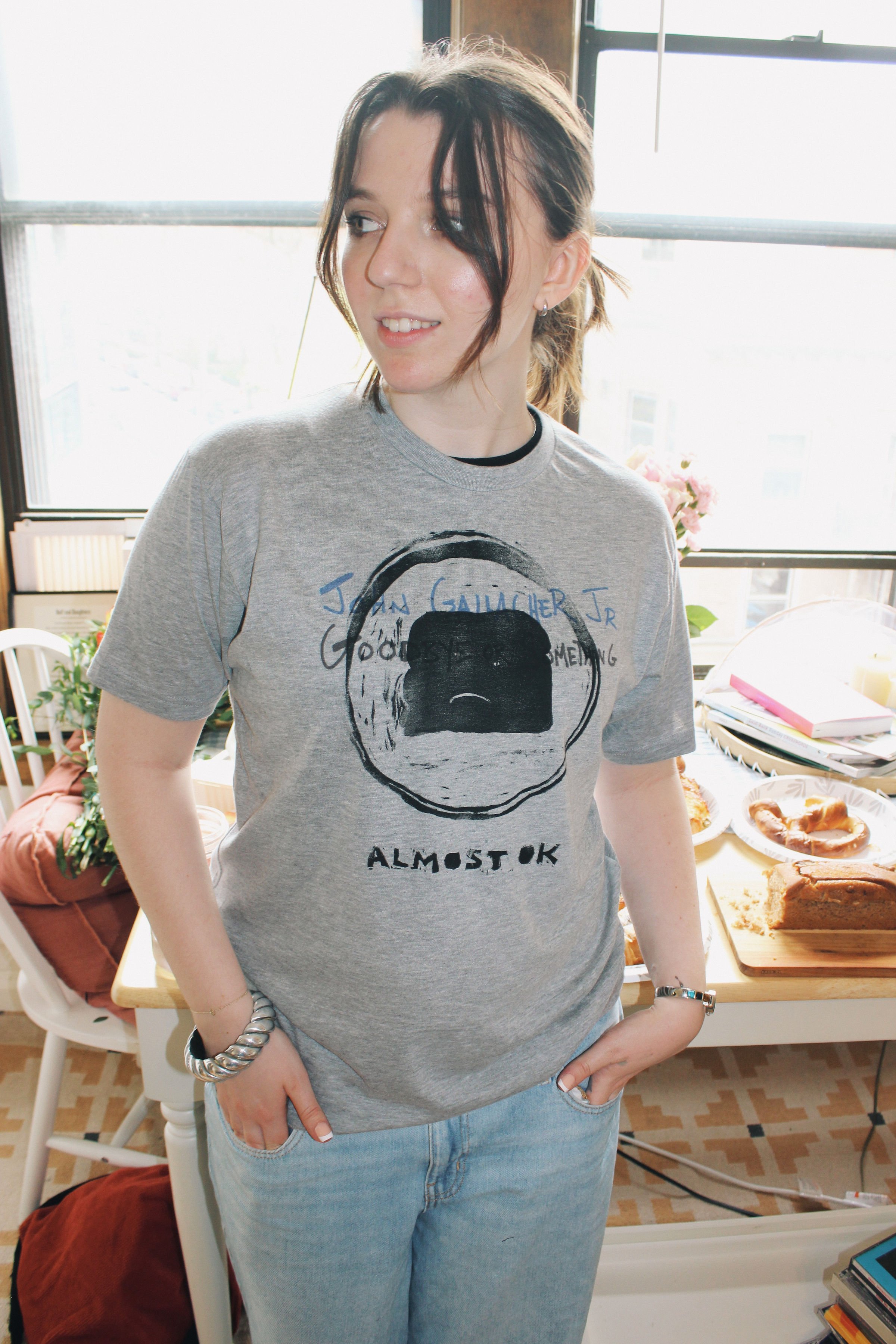 A young woman with short dark hair, wearing a gray T-shirt with a handmade print and light blue jeans, standing indoors near a window during daytime, with a table of baked goods and papers in the background.