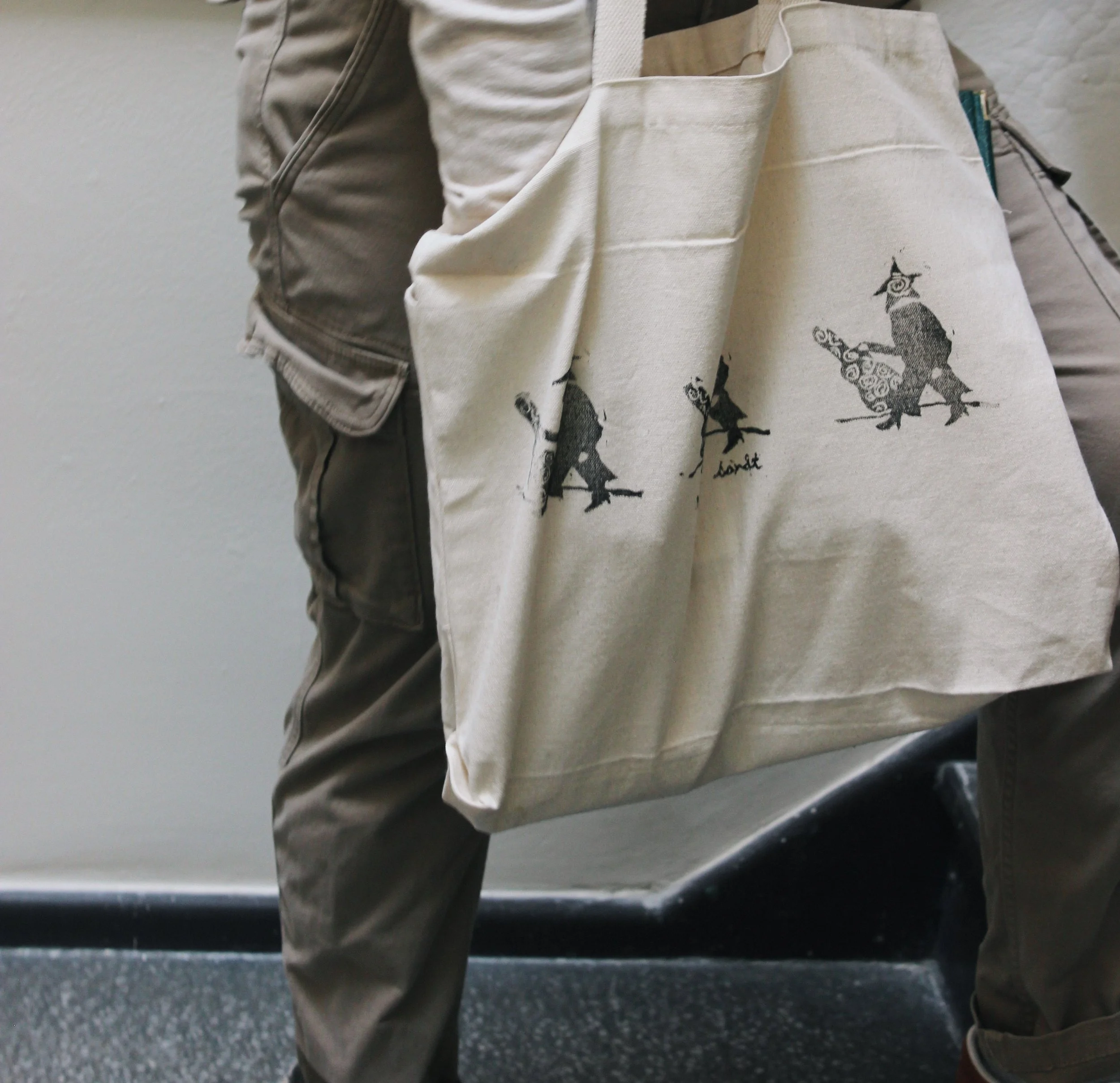 Close-up of a person wearing brown pants and holding a beige tote bag with black artistic illustrations, standing against a gray wall and black floor.