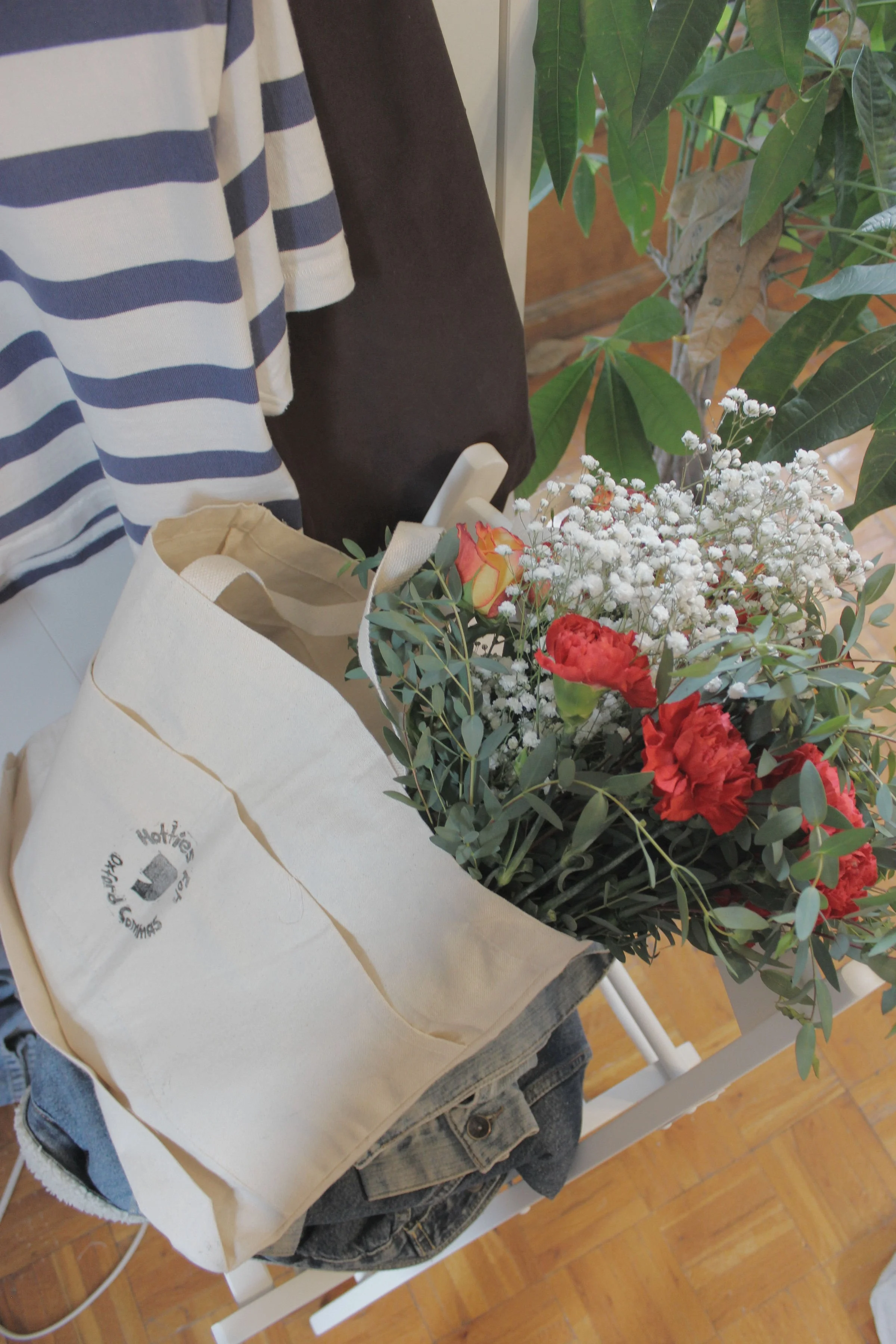 An illustrated shopping bag containing a bouquet of red, orange, and white flowers on a white chair with a pair of blue jeans, in a room with wooden flooring and green leafy plants.