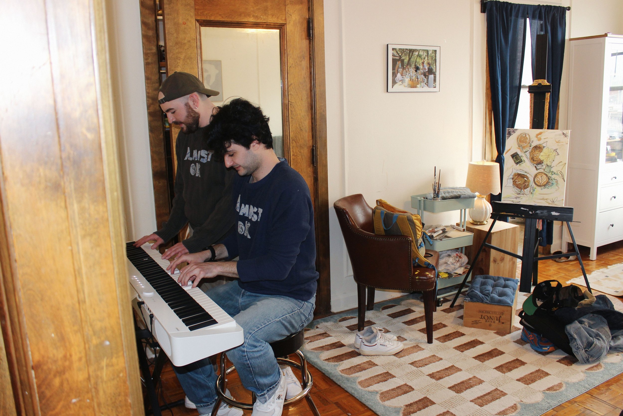 Two young men playing a white electric keyboard in a cozy, cluttered living room. One with black hair, wearing a navy sweatshirt, the other with a beard, wearing a black hoodie and cap, both smiling and focused on the piano.