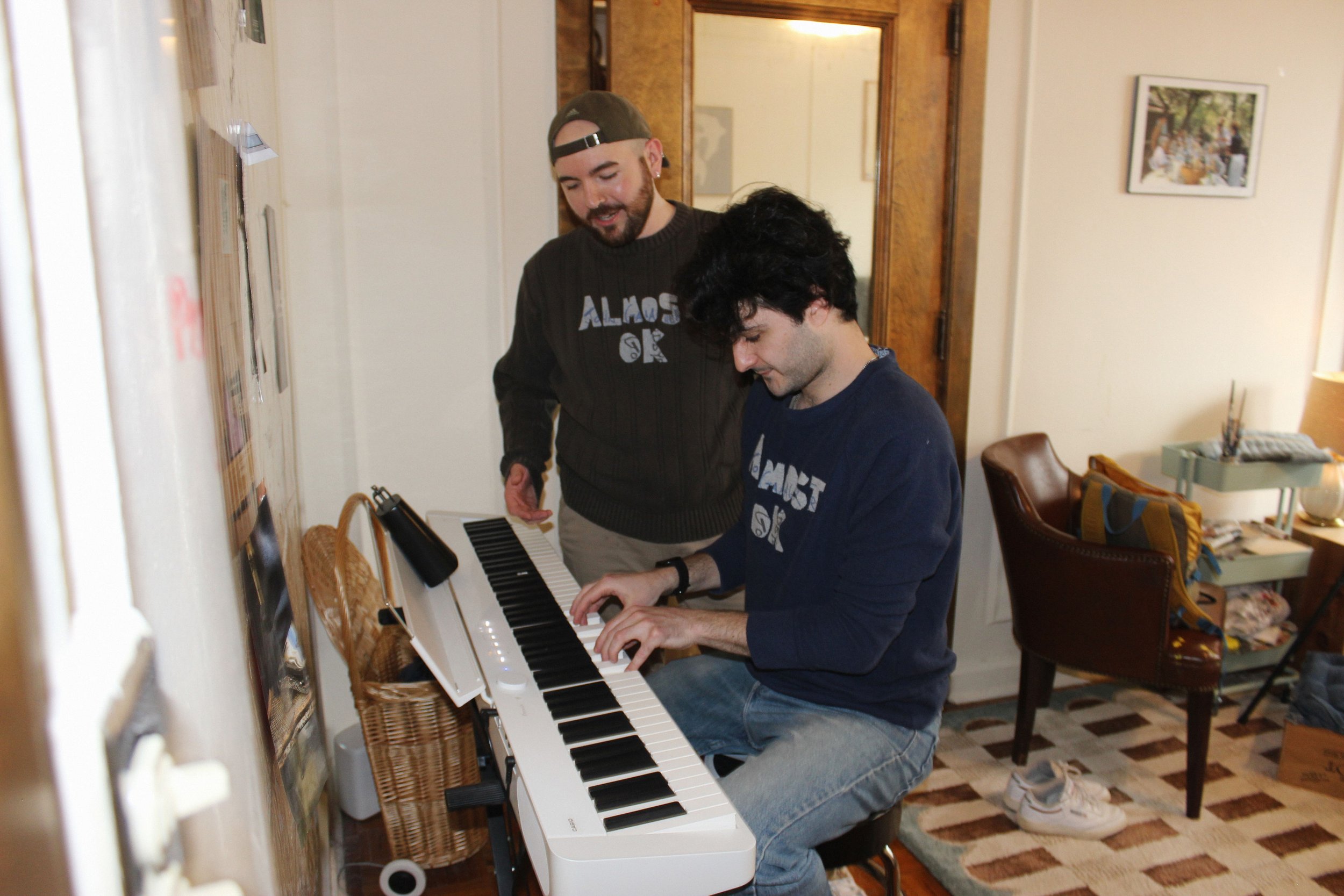 Two men, one sitting at a keyboard and the other standing, in a cozy living room with framed photos on the wall, a brown chair, and a small side table with a yellow bag and lamp. They are wearing sweaters with handmade patches.