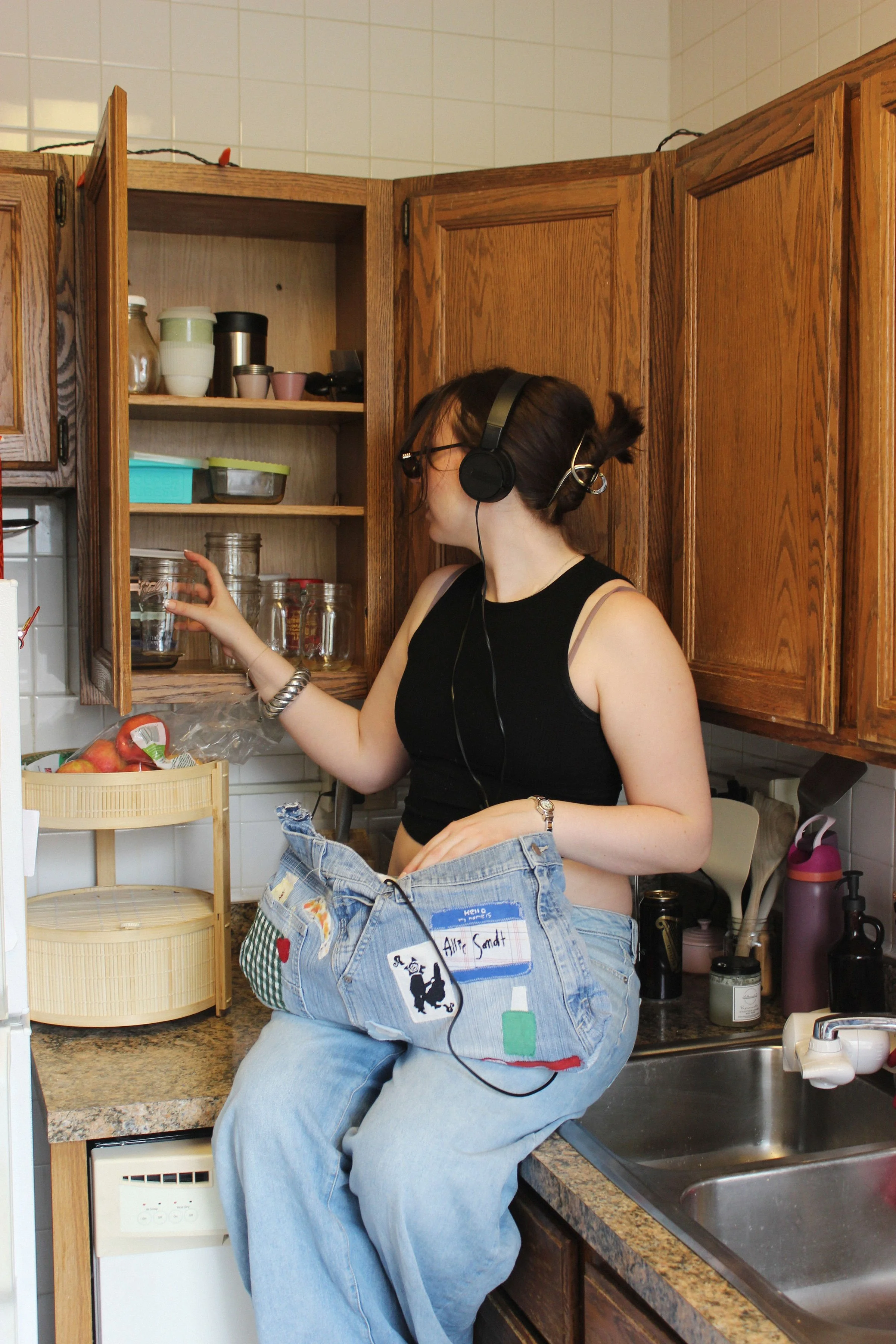 Woman with headphones and glasses sitting on a kitchen counter, reaching into an open cabinet, wearing a black tank top and baggy jeans with handmade patches and a name tag.