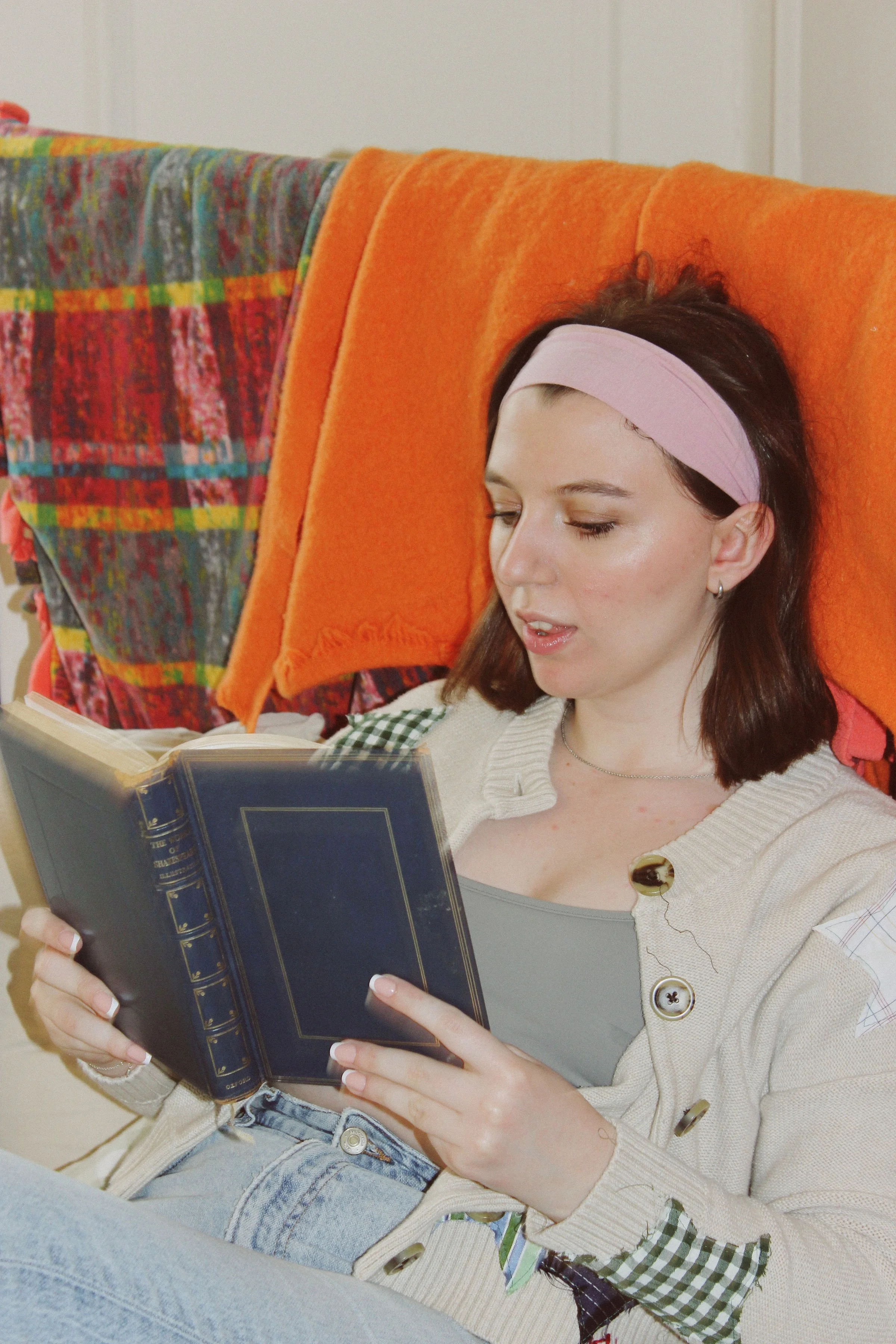 A young woman with a pink headband reading a book while sitting on a couch with colorful and orange blankets.