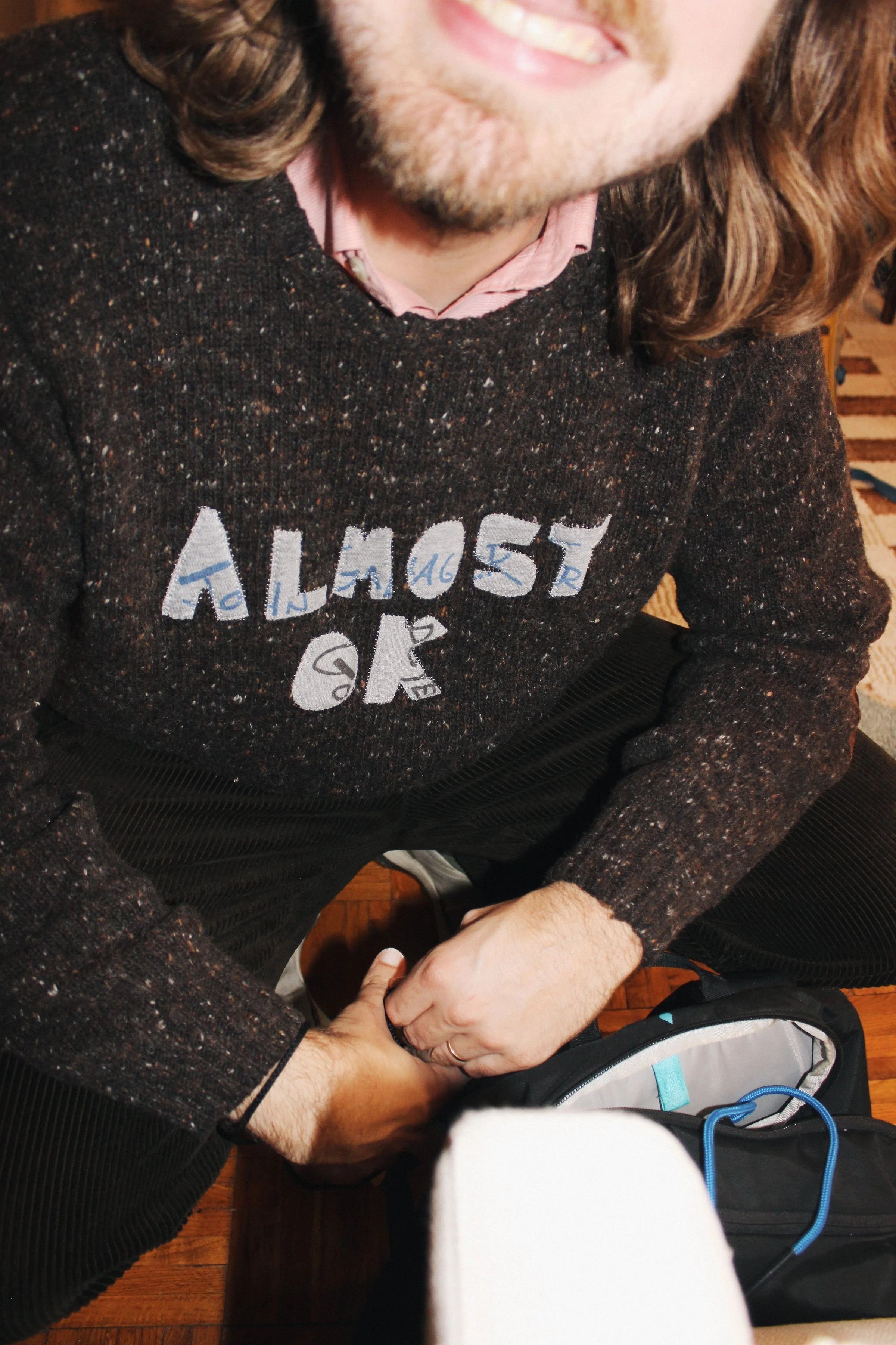 A man with long wavy hair and a beard smiling, wearing a dark sweater with multicolored specks and patches, a pink collared shirt underneath. He is sitting and holding a backpack.