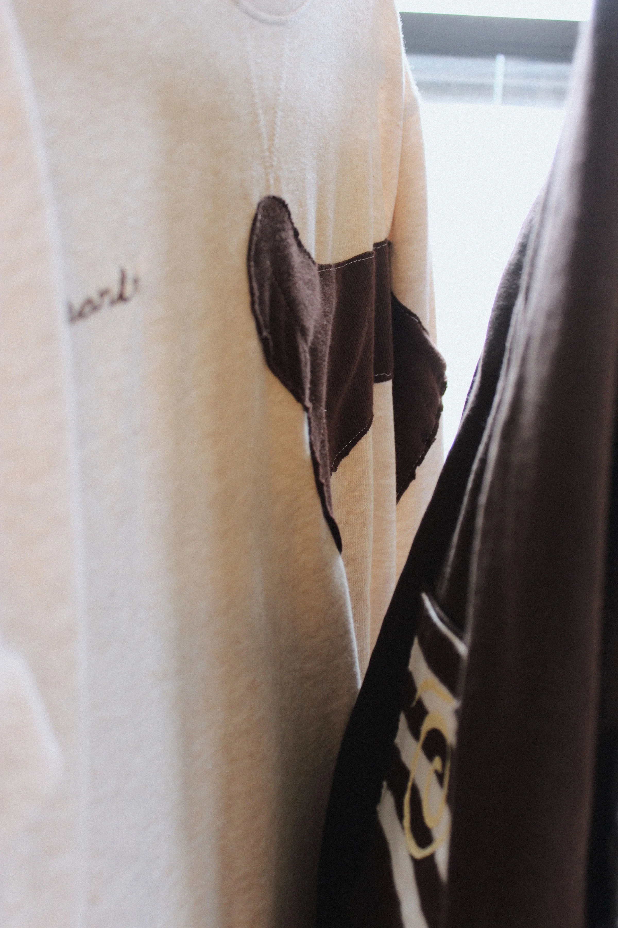 A beige sweater with brown patches on the sleeve and a small embroidered text on the chest, hanging next to a dark jacket with a graphic design.