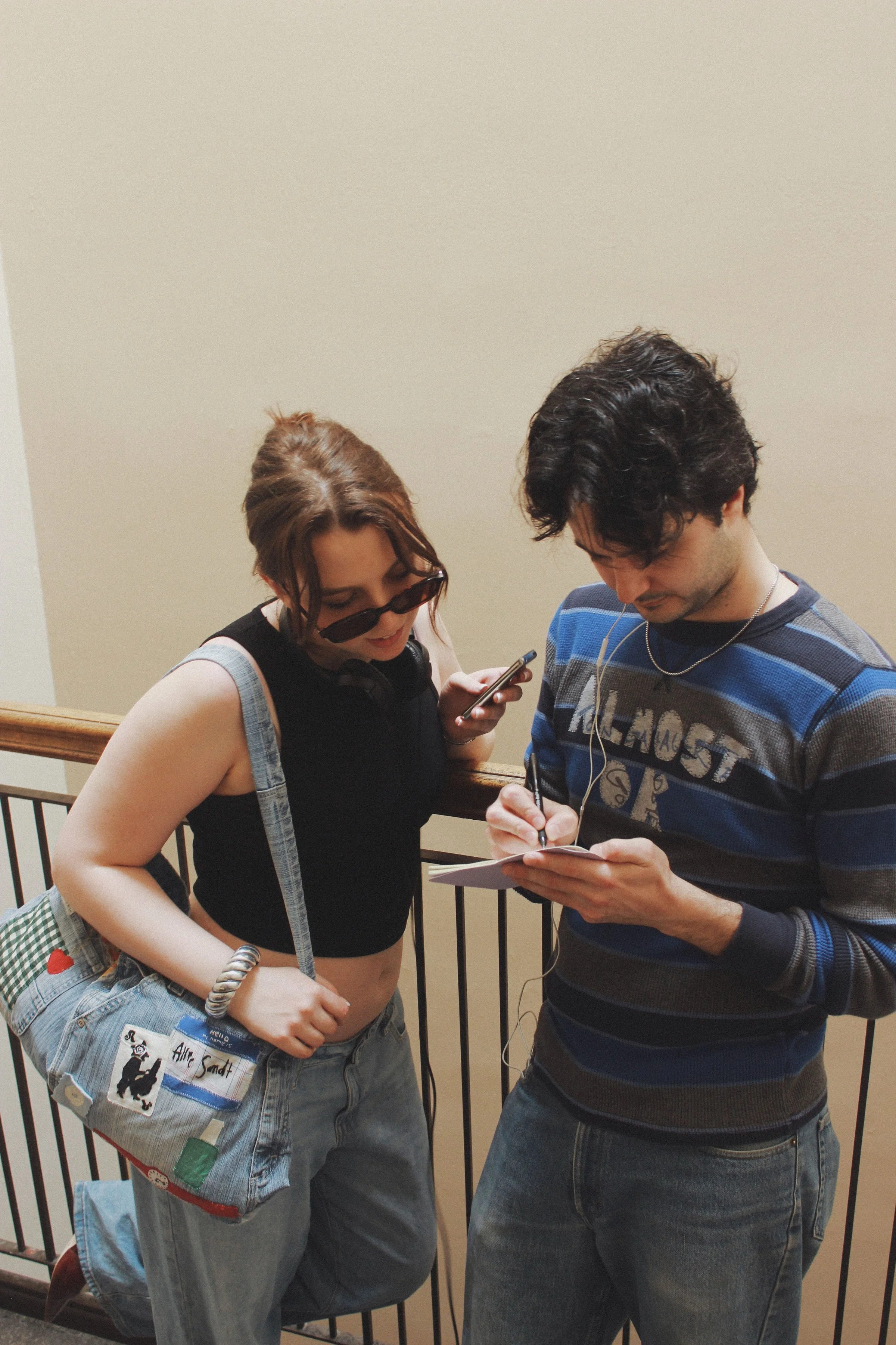 Two young people standing on a staircase, looking at a notebook or phone. The woman has red hair, sunglasses, a black sleeveless top, and a patchwork bag. The man has dark, curly hair, a striped sweater, and is holding a pen. They both wear earphones