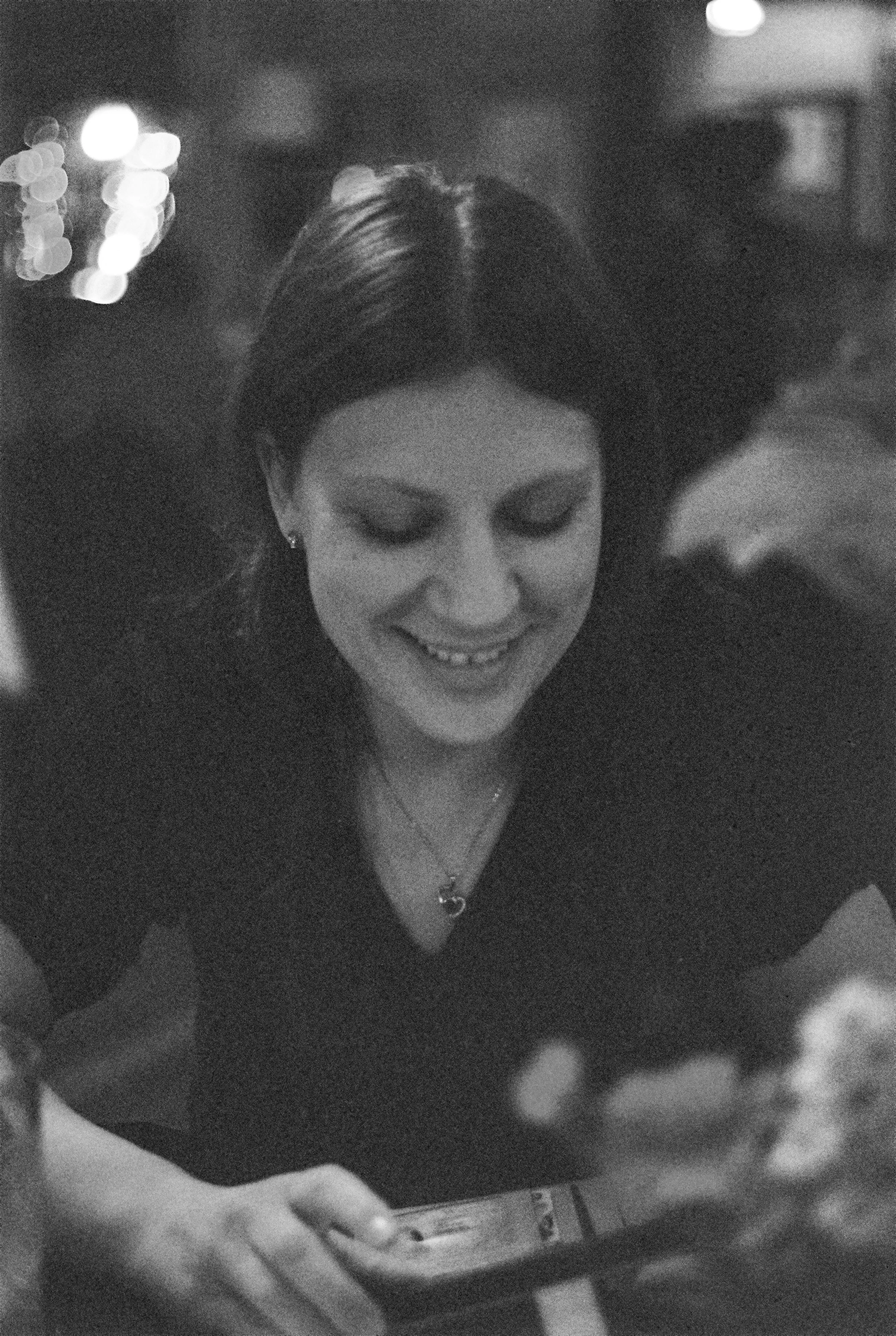 A woman with dark hair and a smile, holding a smartphone and looking at it in a dimly lit room.