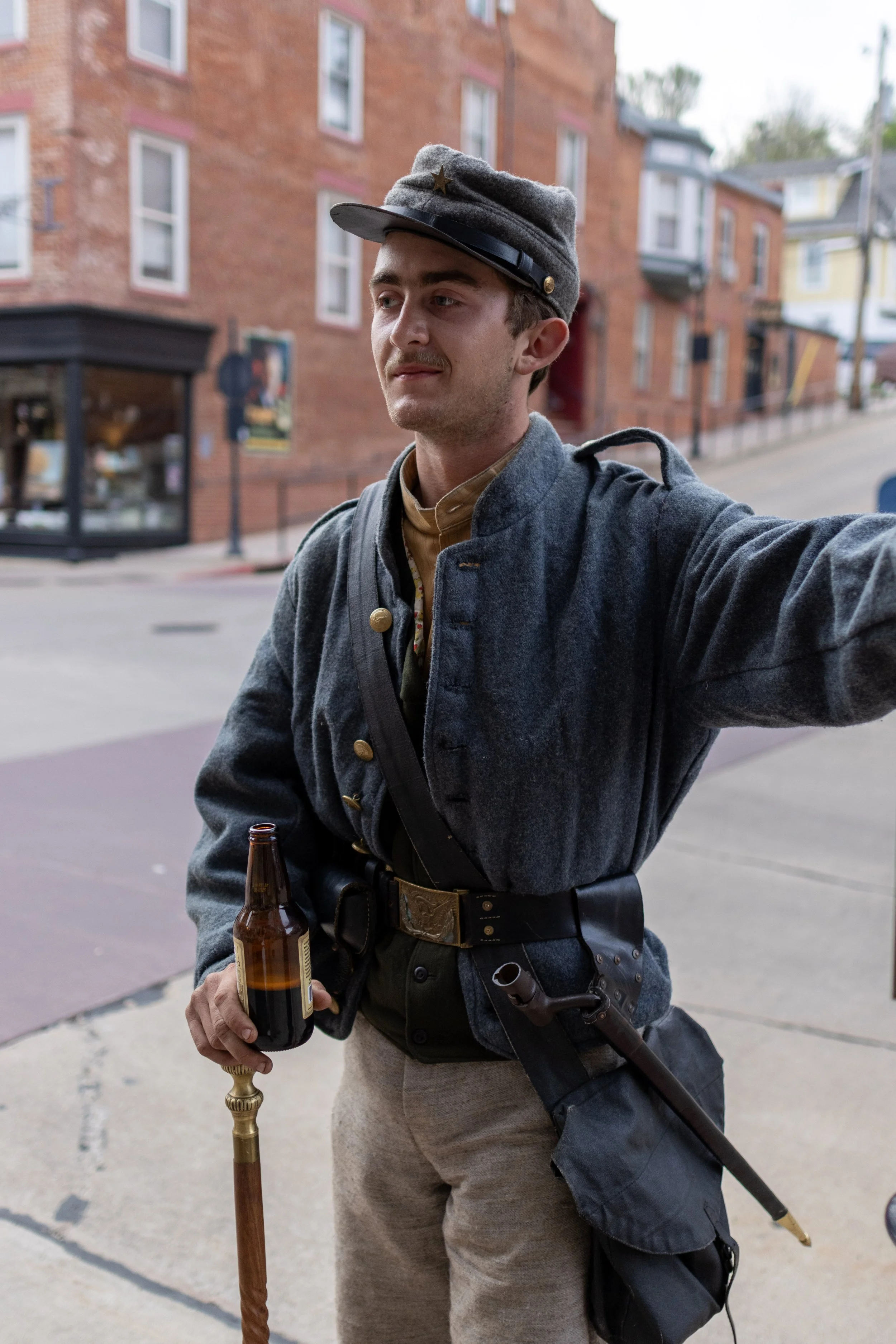 A man dressed in historical military costume, wearing a gray uniform and a cap with a star, holding a beer bottle and a cane, standing outdoors on a city sidewalk with brick buildings in the background.