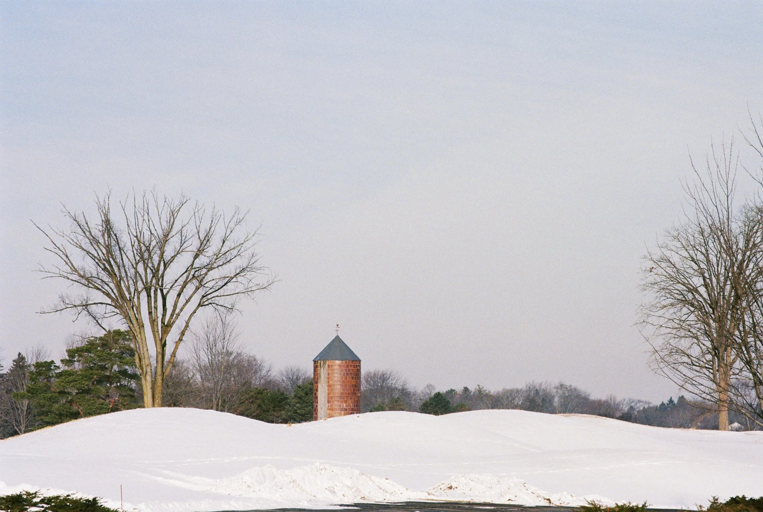 Snow-covered landscape with bare trees, a small brick tower with a conical roof, and an overcast sky.