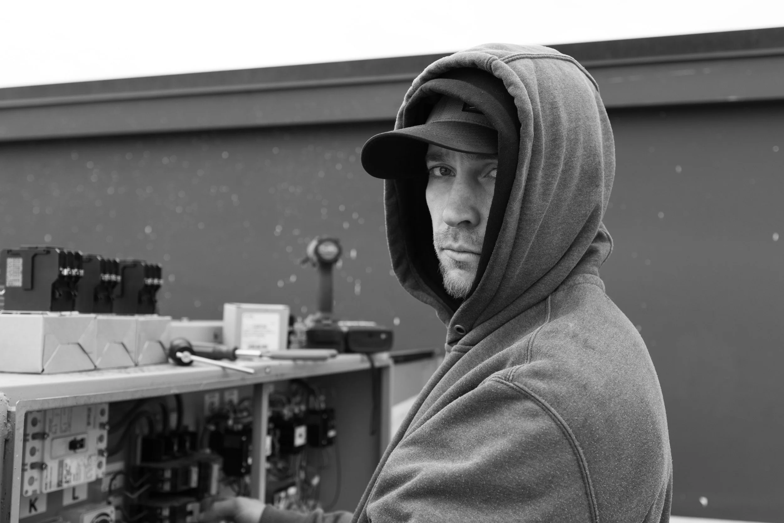 A person wearing a hoodie and cap working on electrical equipment at a workbench.