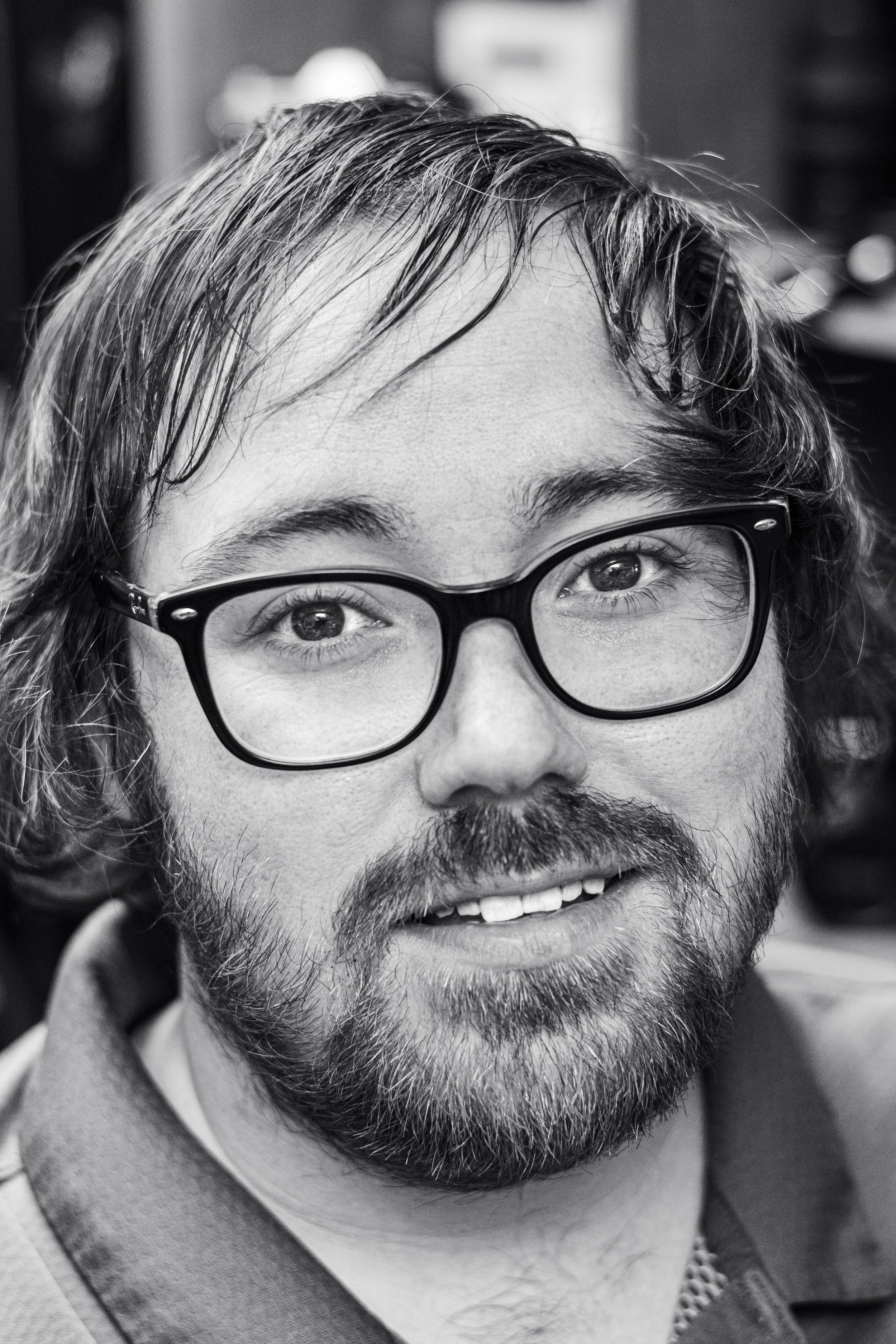 Close-up black and white photo of a person's face, showing their eyes, glasses, hair, nose, mustache, and beard.