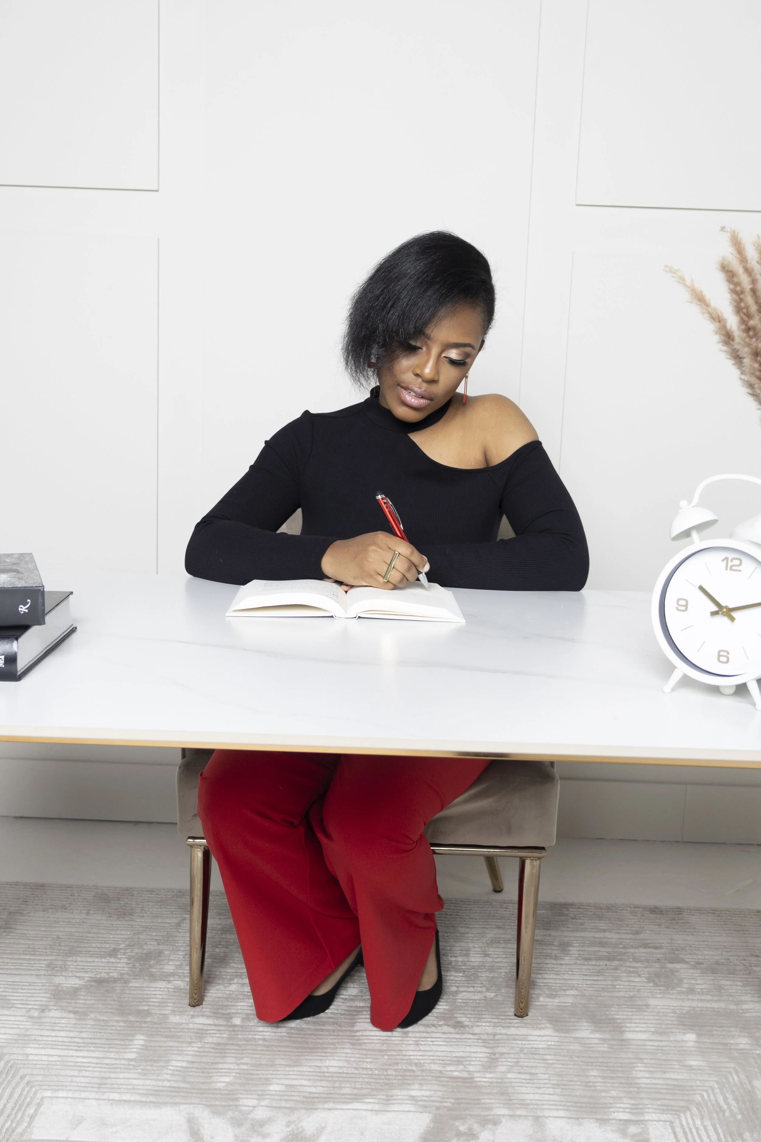A woman sitting at a white desk, writing in a notebook with a red pen. She has black hair, is wearing a black off-shoulder top and red pants, and is seated on a beige chair. A white clock and a small stack of black books are on the desk.