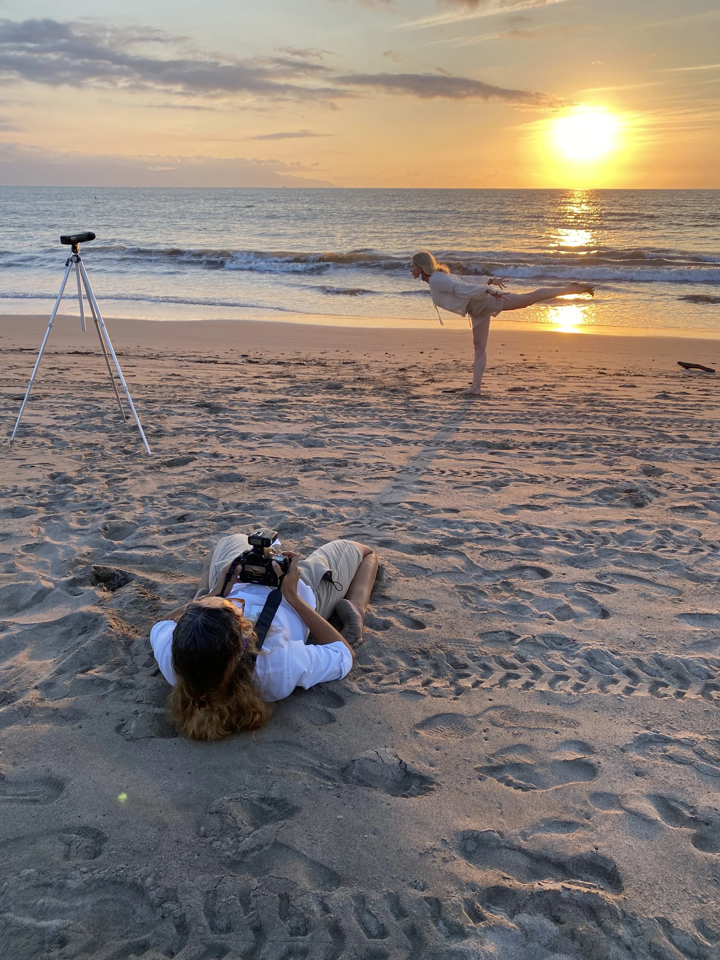 A woman practicing yoga on the beach during sunset with a photographer lying on the sand capturing her pose, a camera on her chest, and a telescope set up nearby.