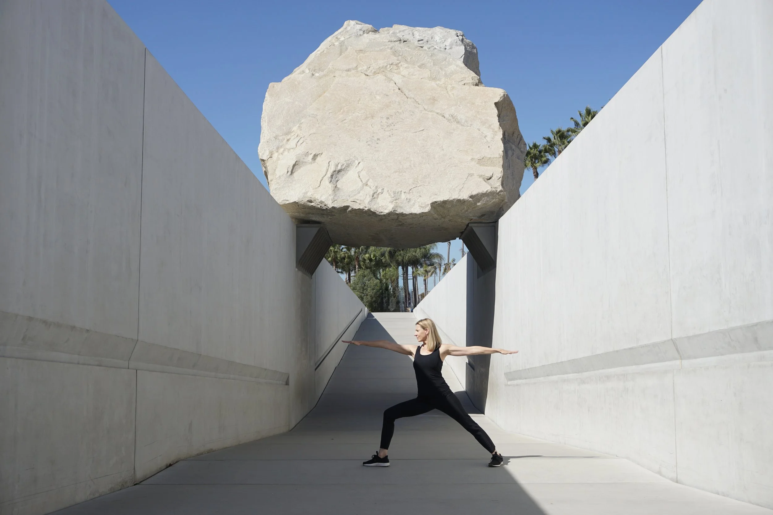 A woman in black athletic wear performing warrior yoga pose underneath a large hanging rock between two concrete walls, with palm trees and blue sky in the background.