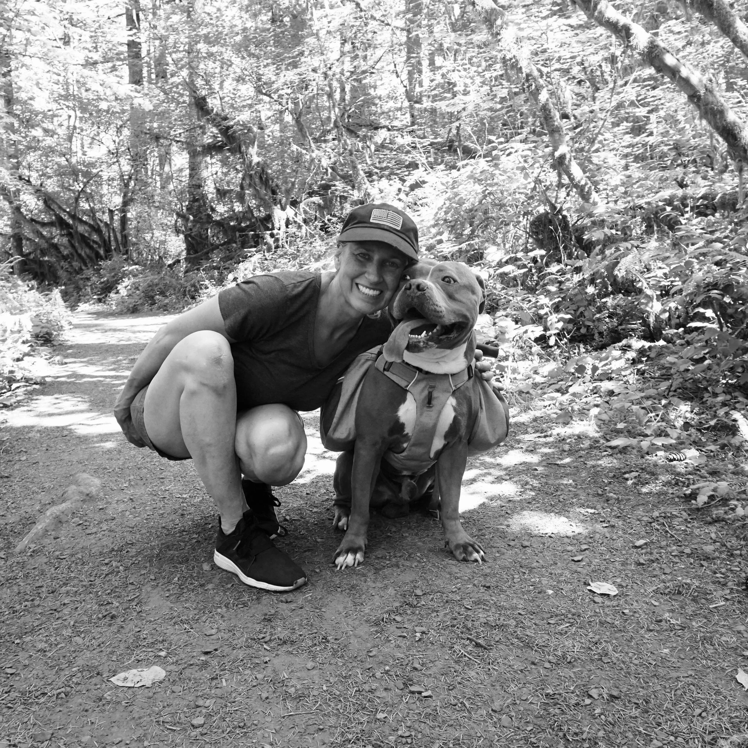 A woman and a large dog with a backpack sitting on a trail in a forest.