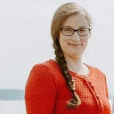 Allison Outlund, a woman in a red knitted sweater, smiling and standing outdoors in natural light in a professional portrait