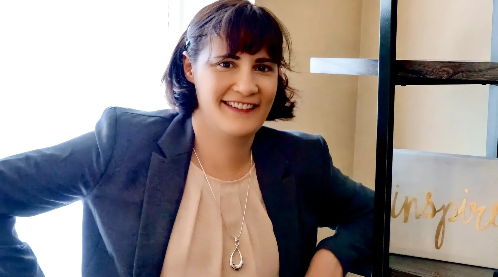 Olivia Outlund, a woman in a blazer, smiling and leaning on a shelf in a professional office setting