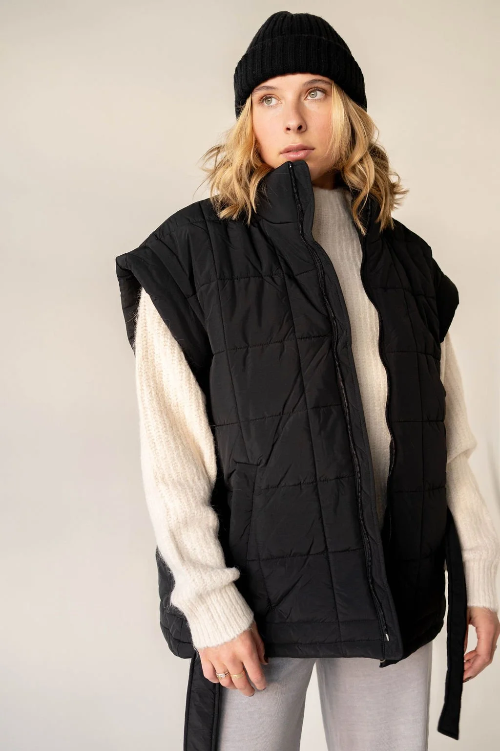A woman wearing a black knit beanie, a black puffer vest, a beige sweater, and gray sweatpants, standing against a plain background.
