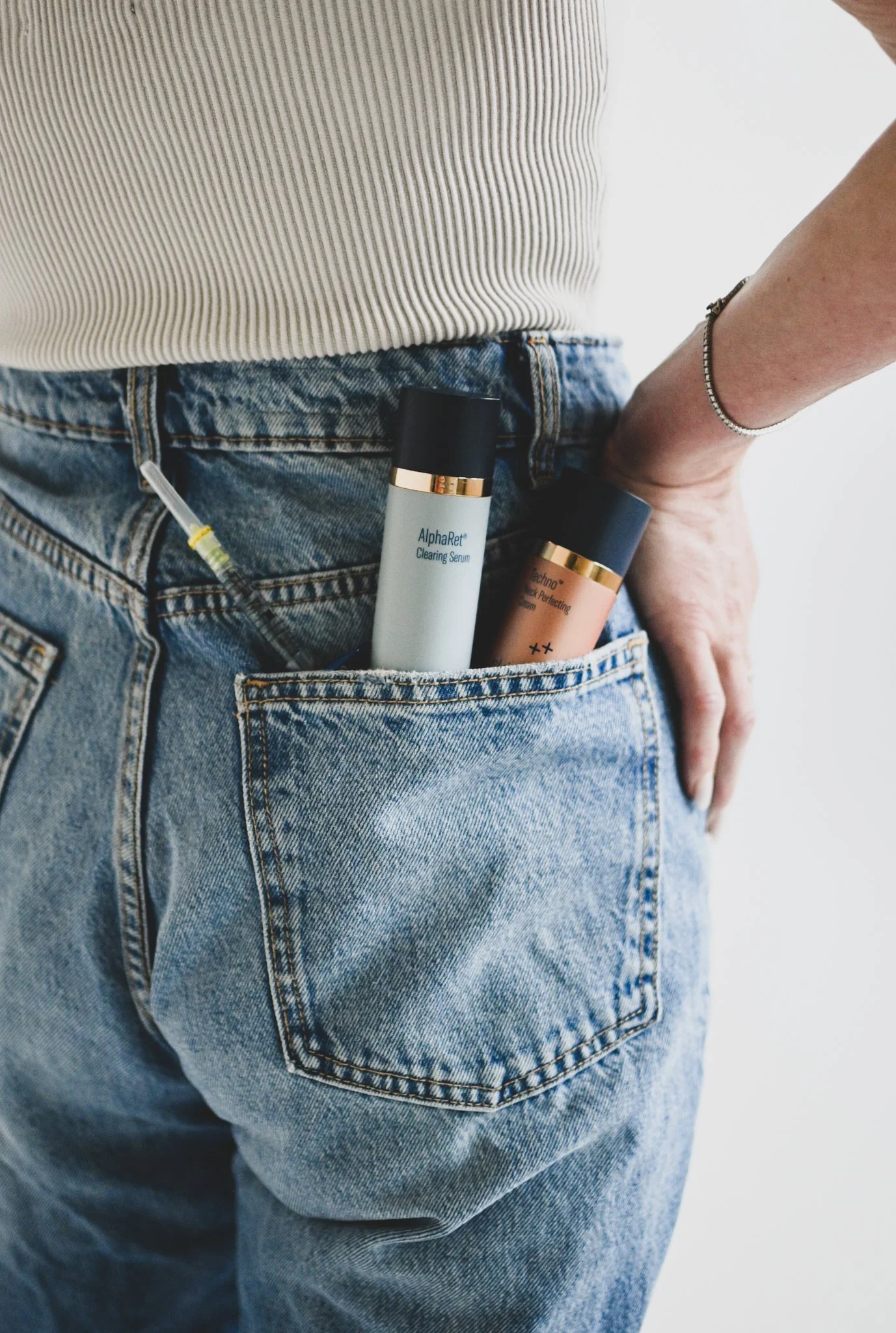 Back pocket of blue jeans with skincare products and a syringe inside, partially covered by a person's hand and white striped shirt.
