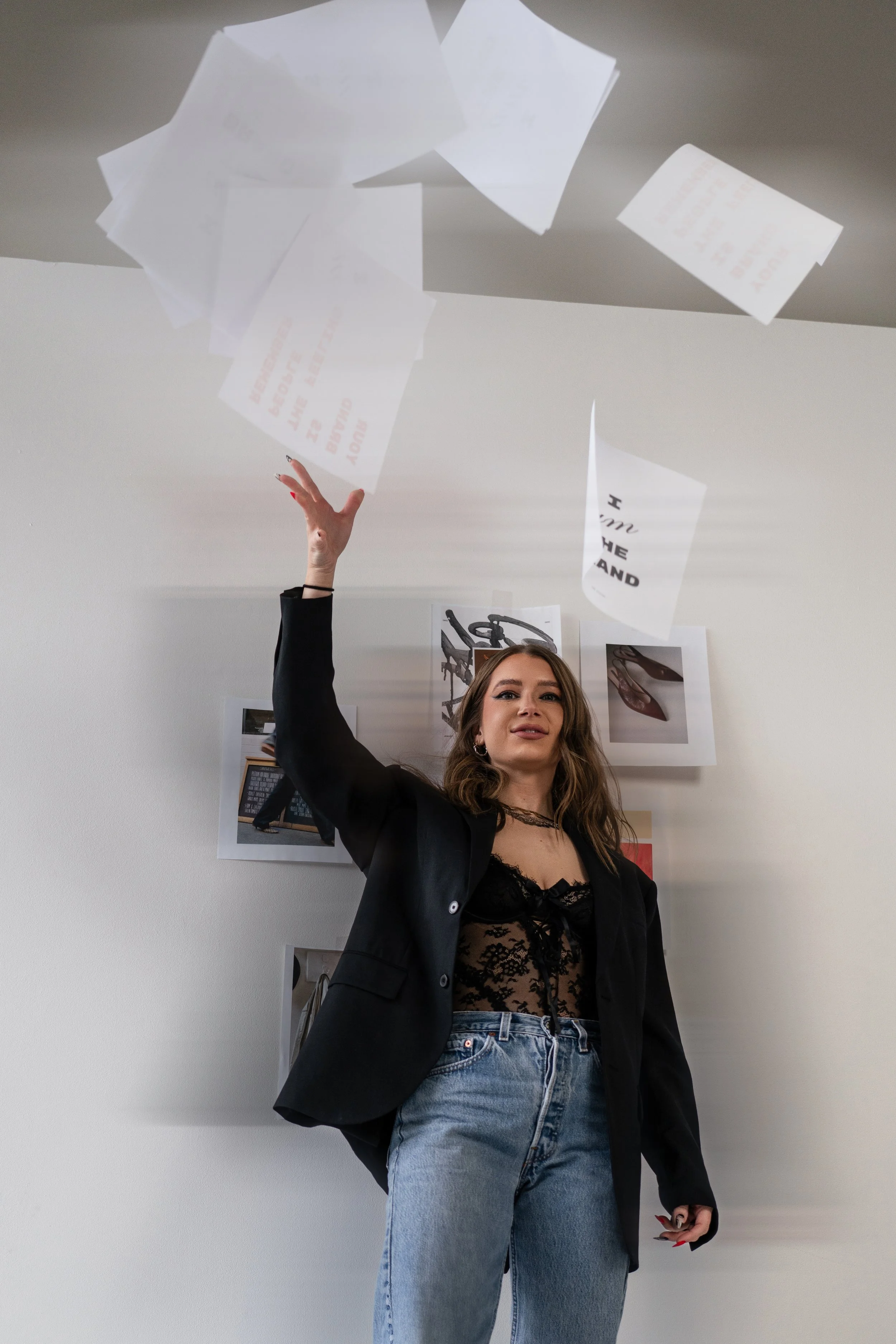 A young woman with wavy brown hair wearing a black lace top, a black blazer, and blue jeans, is tossing papers into the air inside a room with white walls and photos and artwork on the wall behind her.