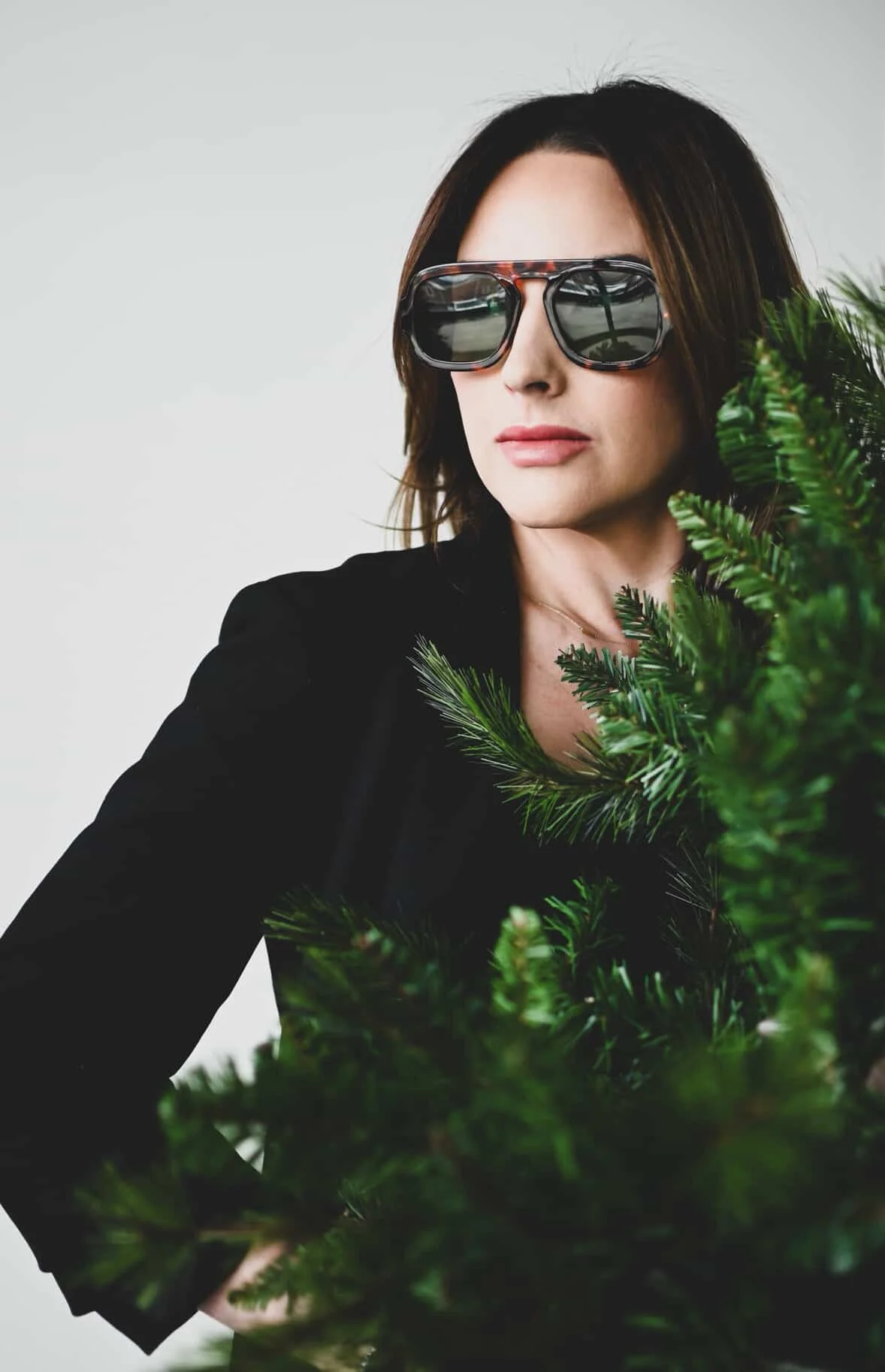 A woman wearing sunglasses and a black top, standing behind a green Christmas tree. She has shoulder-length brown hair and is looking to the side.
