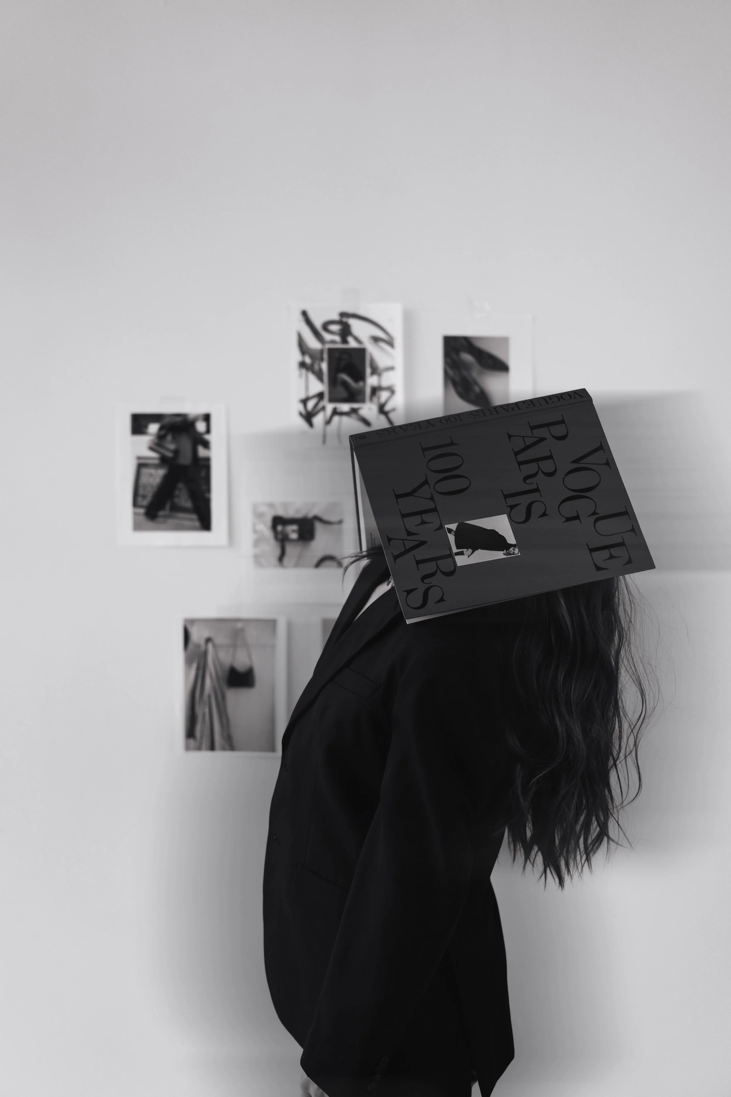A person with long hair wearing a dark jacket is standing against a wall with photos, with a Vogue book titled '100 Years' obscuring their face.