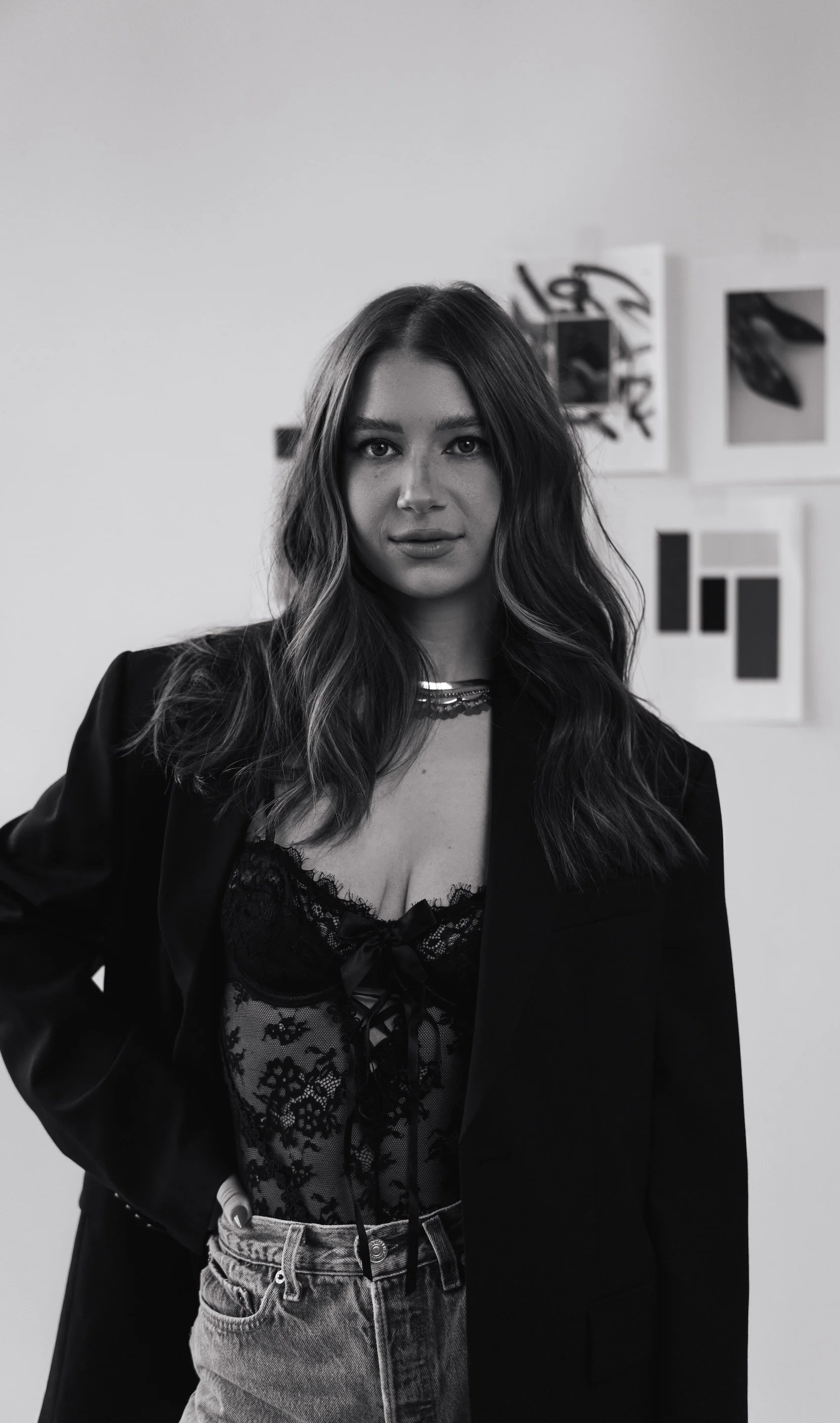 Black and white portrait of a young woman with wavy hair, wearing a lace top, a blazer, and jeans, standing indoors against a white wall with abstract art.