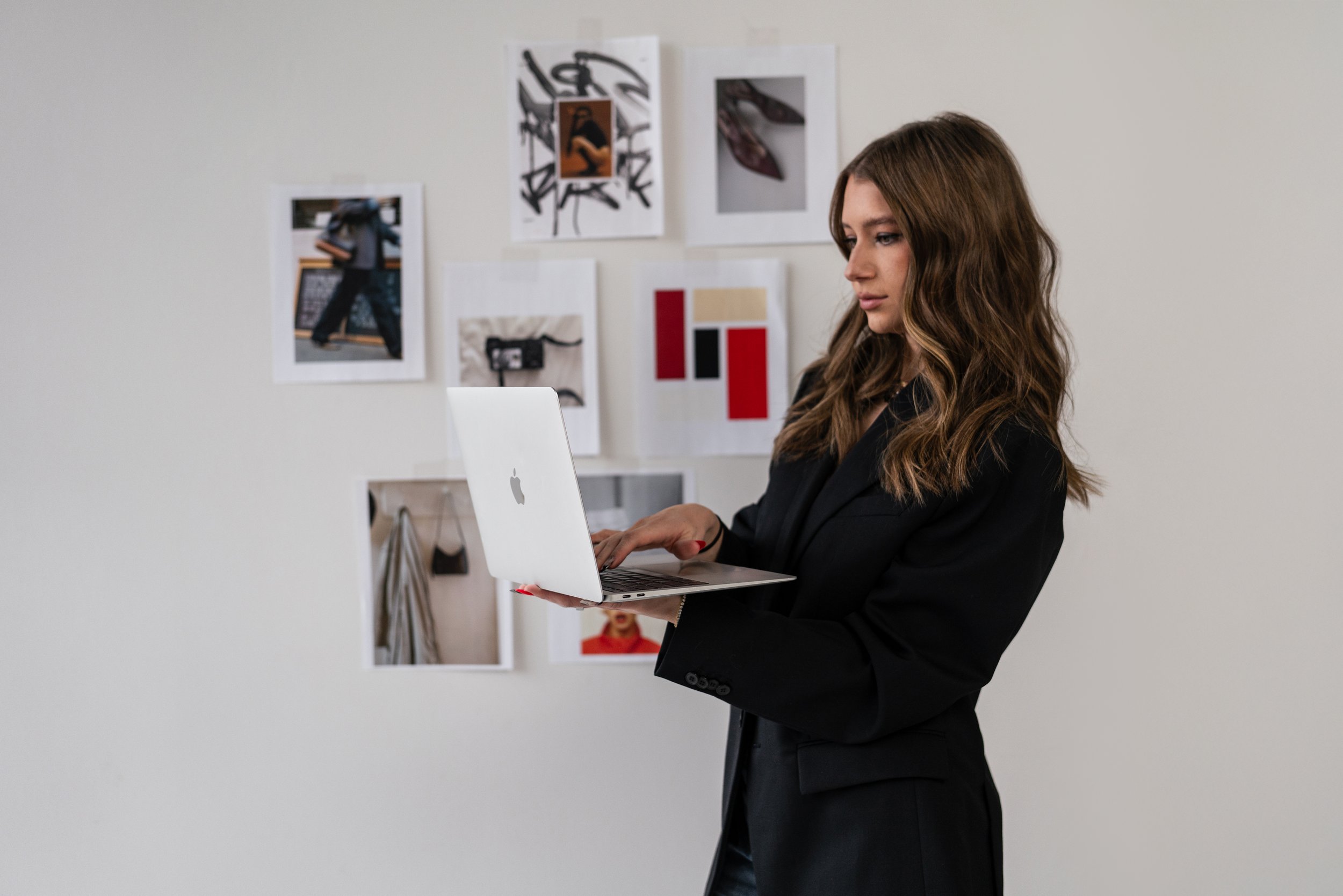 Woman with wavy brown hair wearing a black blazer using a silver Apple MacBook in a room with white walls and framed photos and artwork.