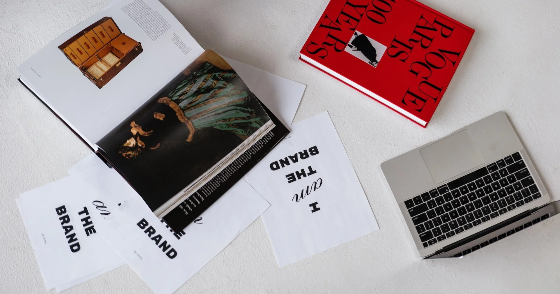 A desk with an open magazine showing interior design images, black and white papers with text, a red book titled 'Vogue Paris', and a silver laptop with a black keyboard.