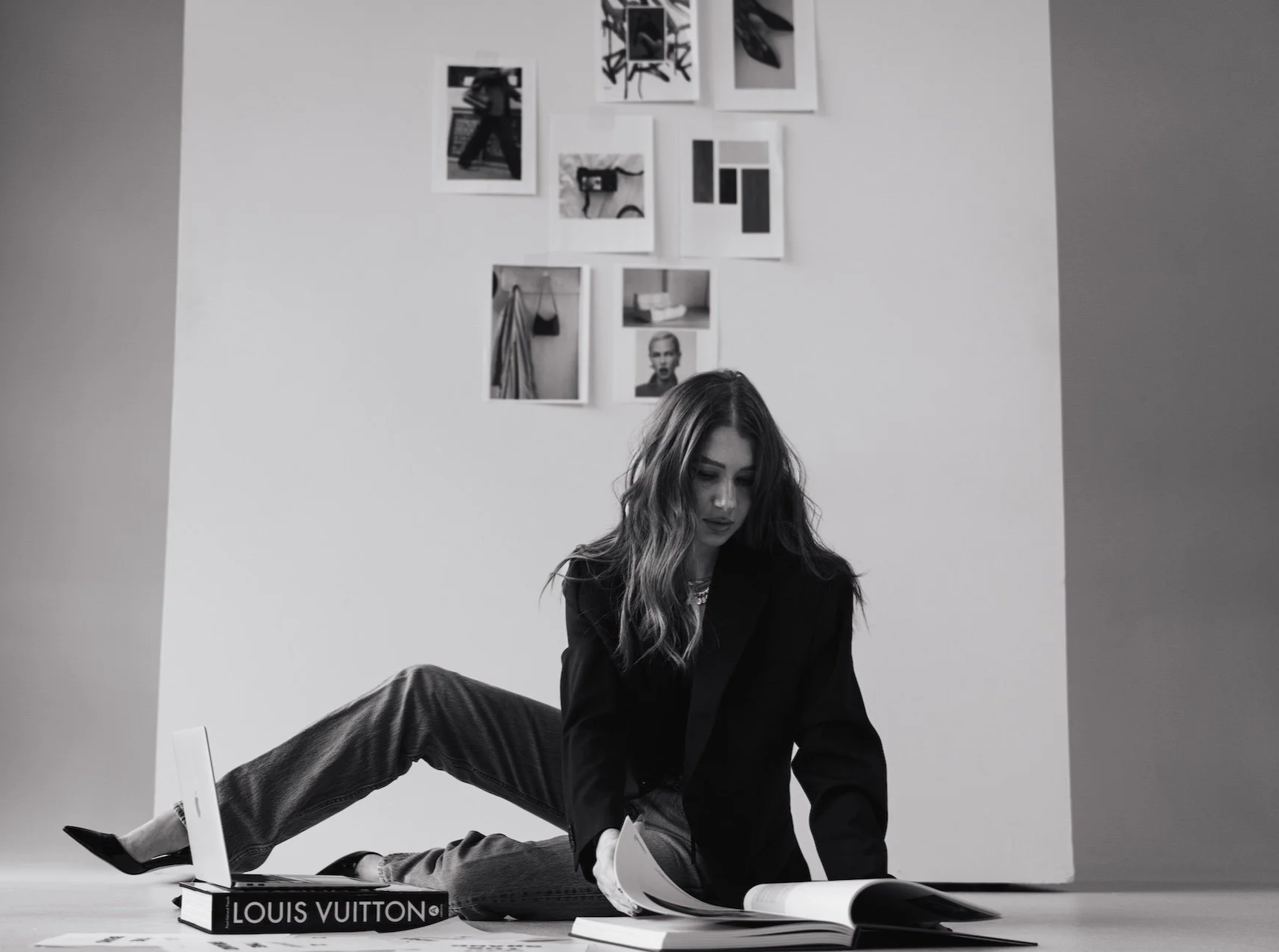 A woman with wavy hair, wearing a blazer and jeans, is sitting on the floor flipping through a large book. There are other books and papers scattered around her, with a Louis Vuitton box nearby. The background features a white wall decorated with several black-and-white photographs.