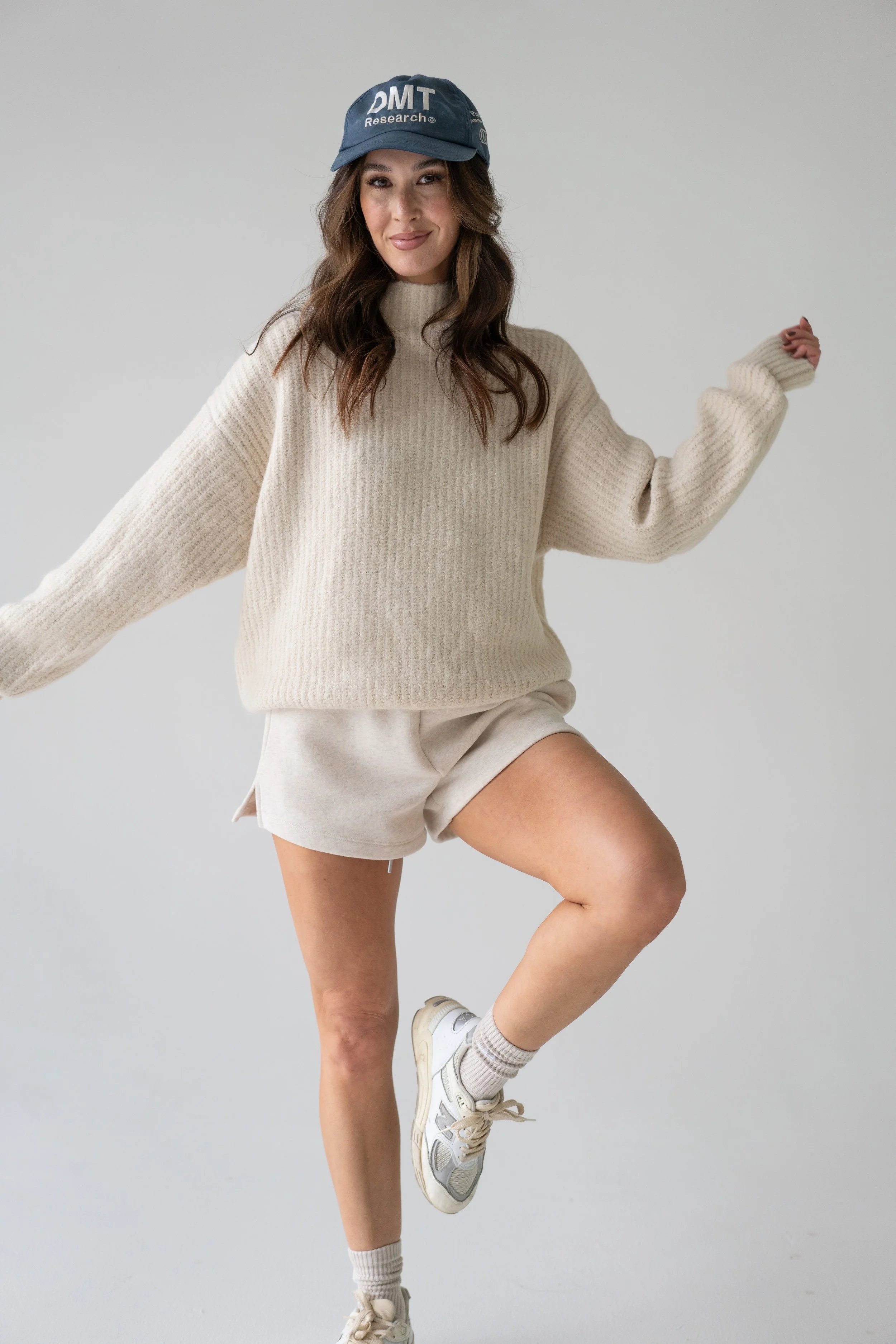Woman wearing a cream oversized sweater, beige shorts, white sneakers, and a blue baseball cap, posing against a plain light background.