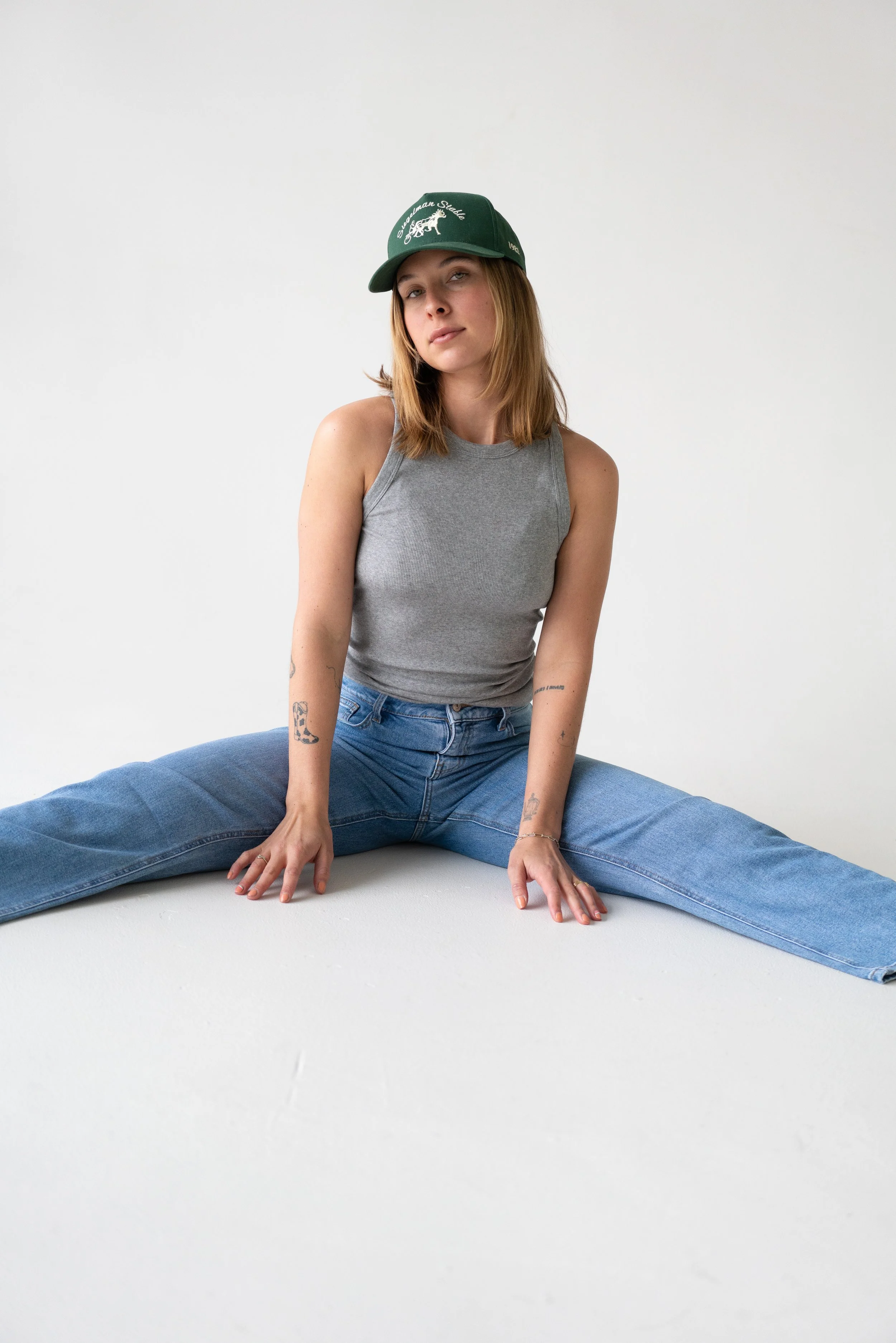 A young woman with tattoos on her arms is sitting on the floor with her legs spread apart, wearing a gray tank top, blue jeans, and a green cap with white embroidery, against a plain white background.