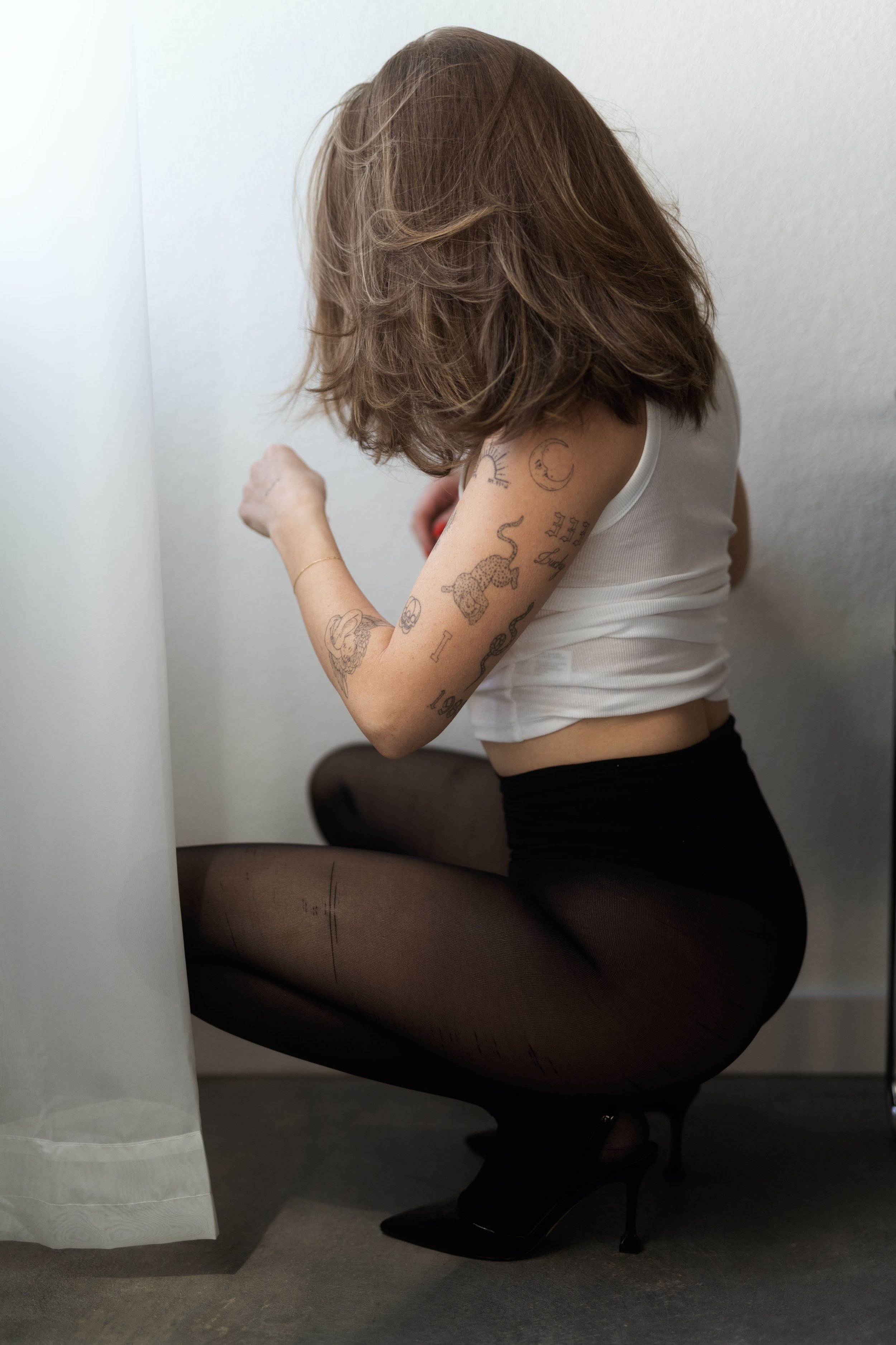 A woman with shoulder-length hair dressed in a white sleeveless top, black tights with ripped details, and black high heels, crouching with her head down, inside a room.