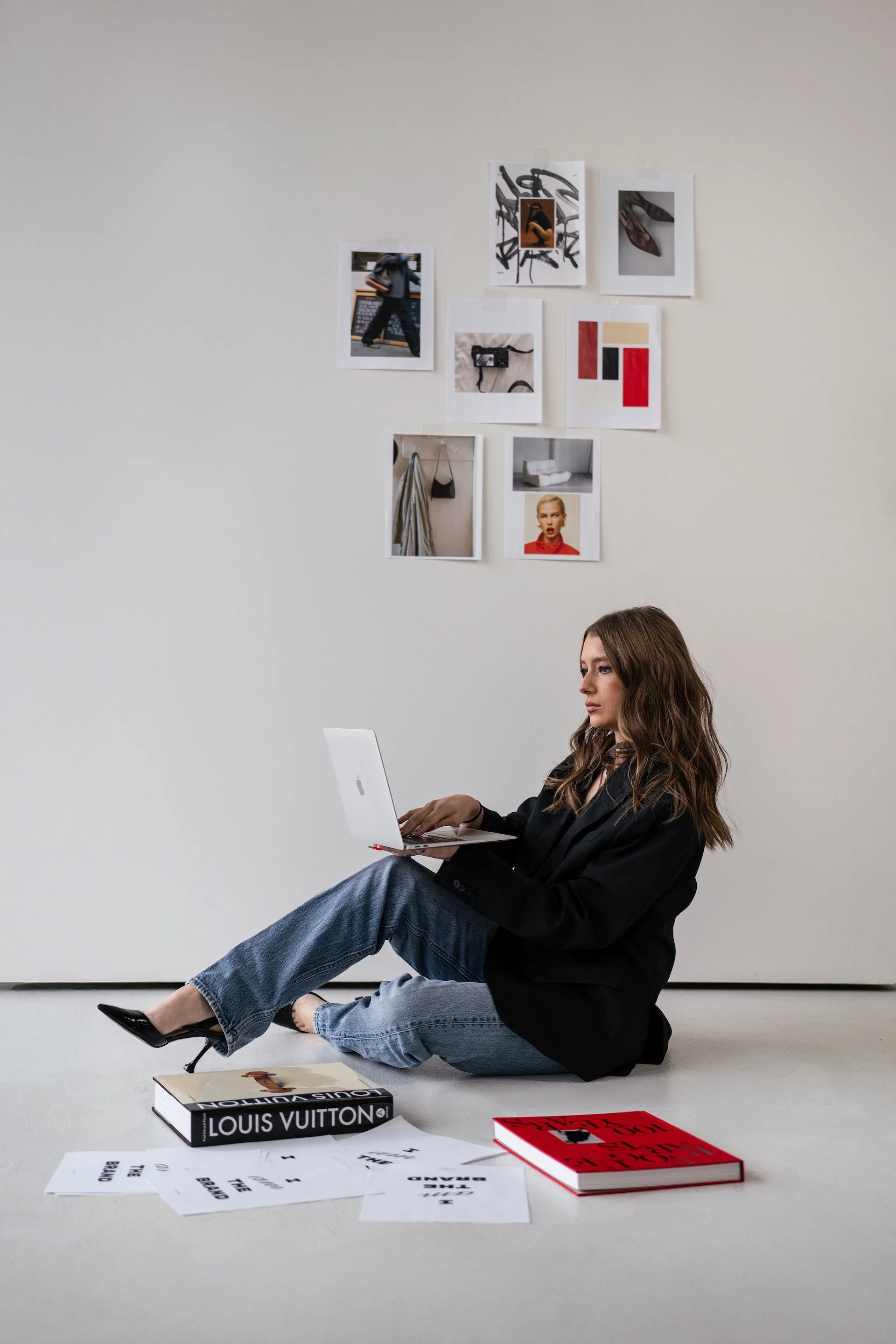 A woman sitting on the floor with a laptop, there are books and papers around her, and photos on the wall behind her.