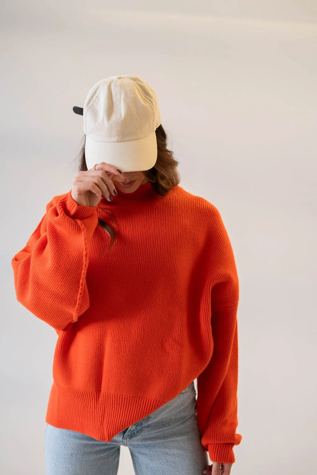 A woman wearing an orange sweater, light jeans, and a beige baseball cap, leaning her head down and holding the cap with her hand, against a neutral background.