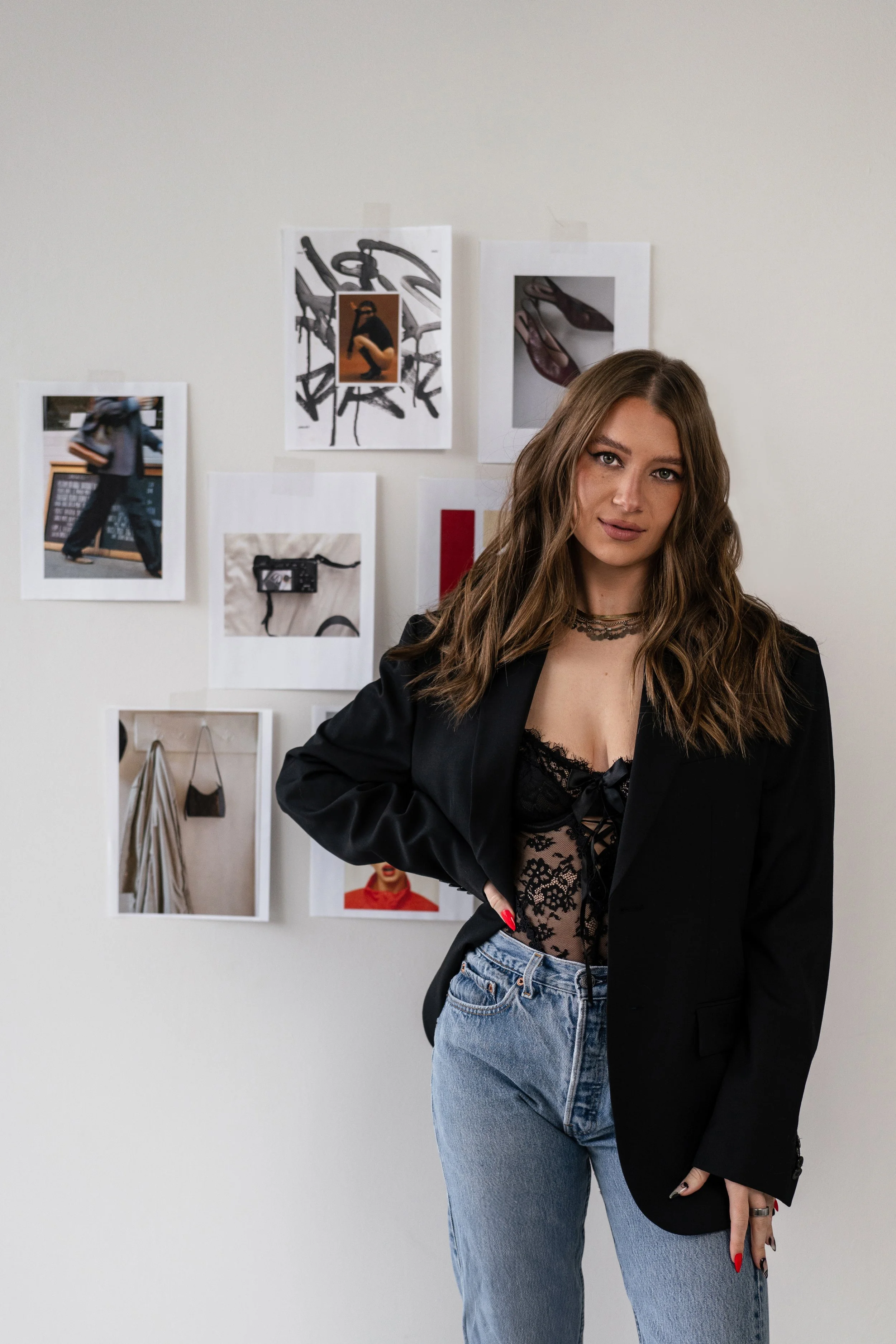 A woman with long wavy brown hair wearing a black lace top, black blazer, and light blue jeans standing in front of a white wall decorated with various photos and artwork.