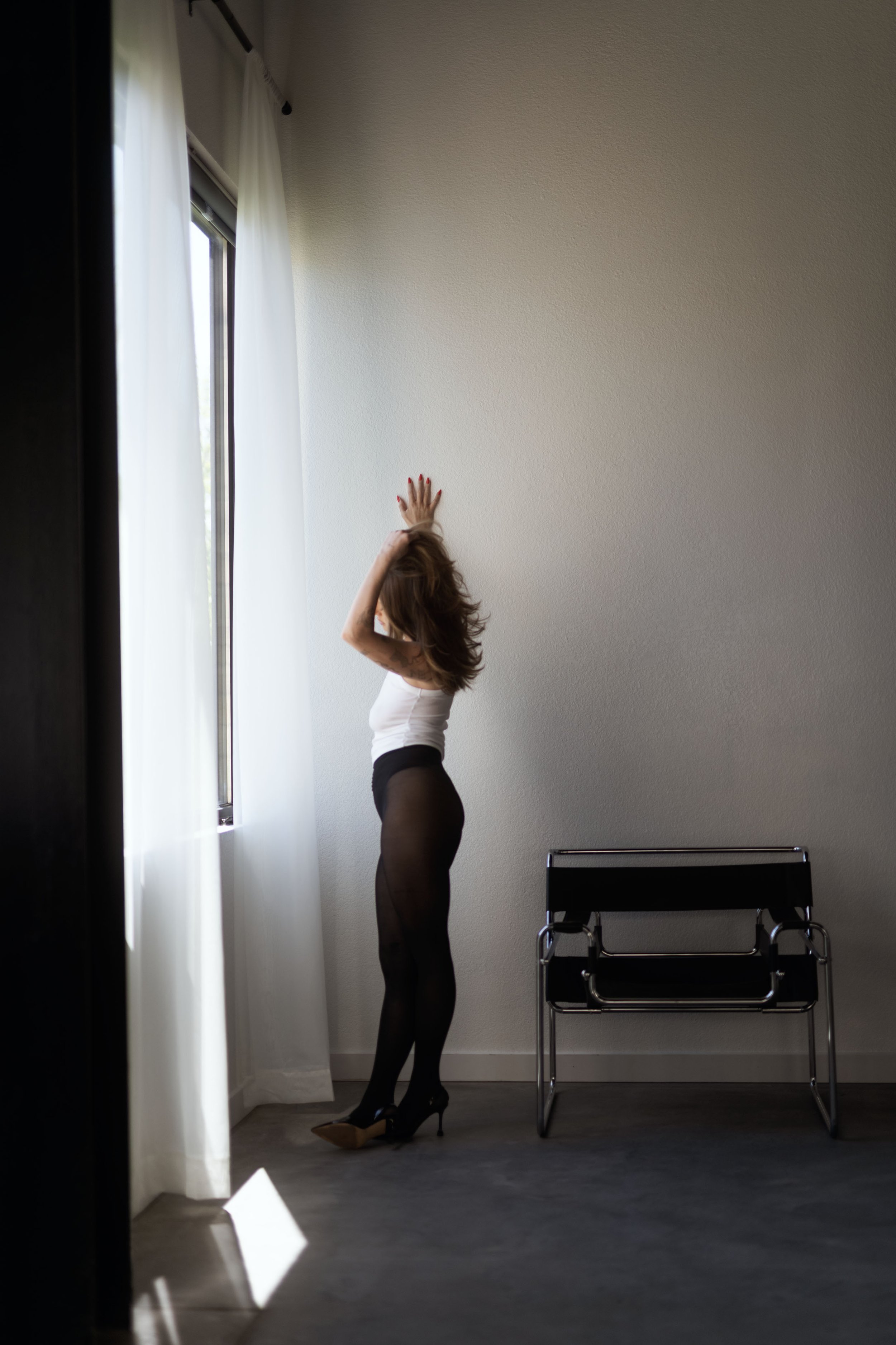 A woman standing near a window with white curtains, dressed in black tights, high heels, and a white top, stretching her arms.