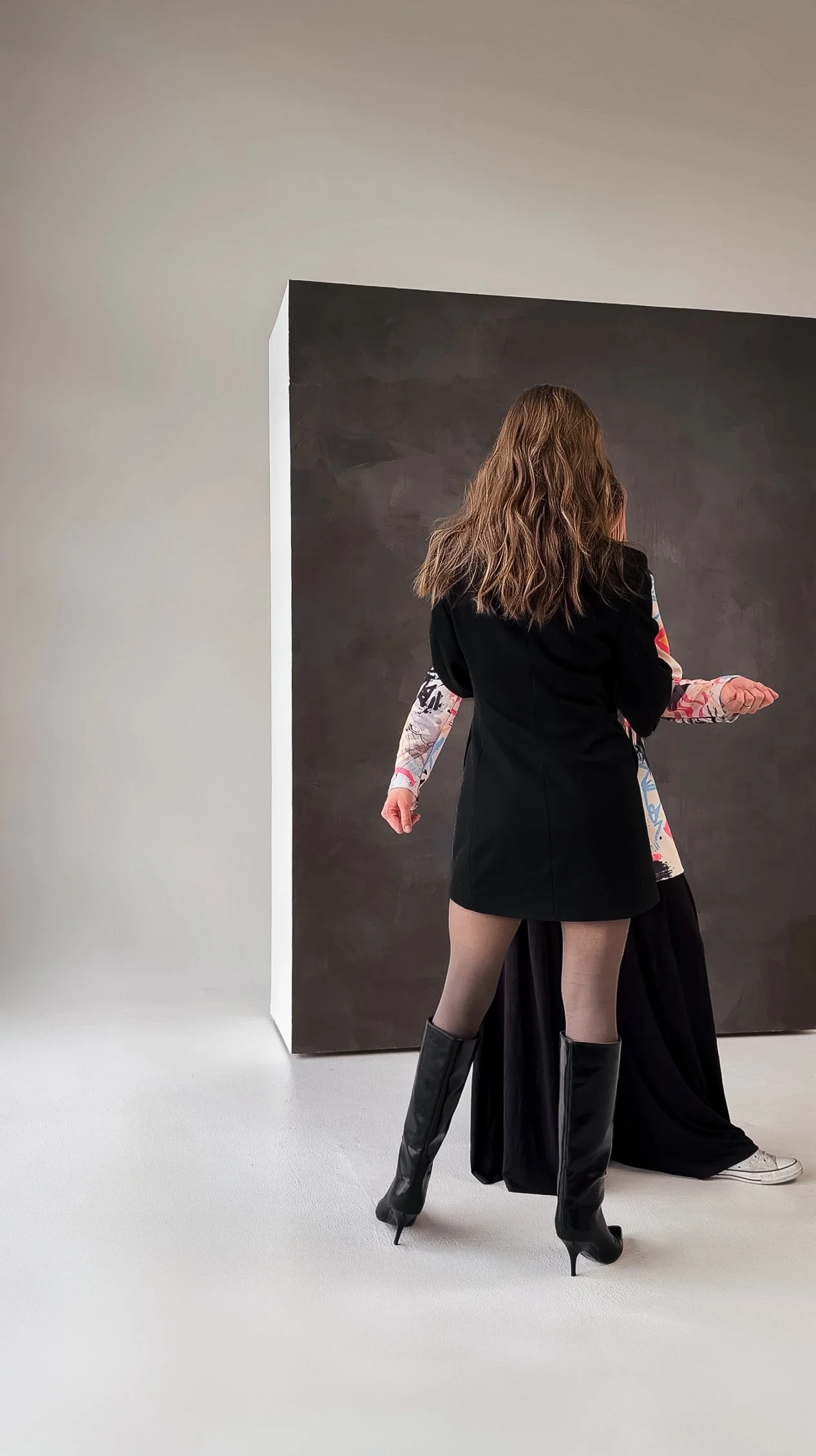 Two women standing together in an art gallery; one is wearing a black dress with knee-high boots, the other has a colorful top with long sleeves and black pants.