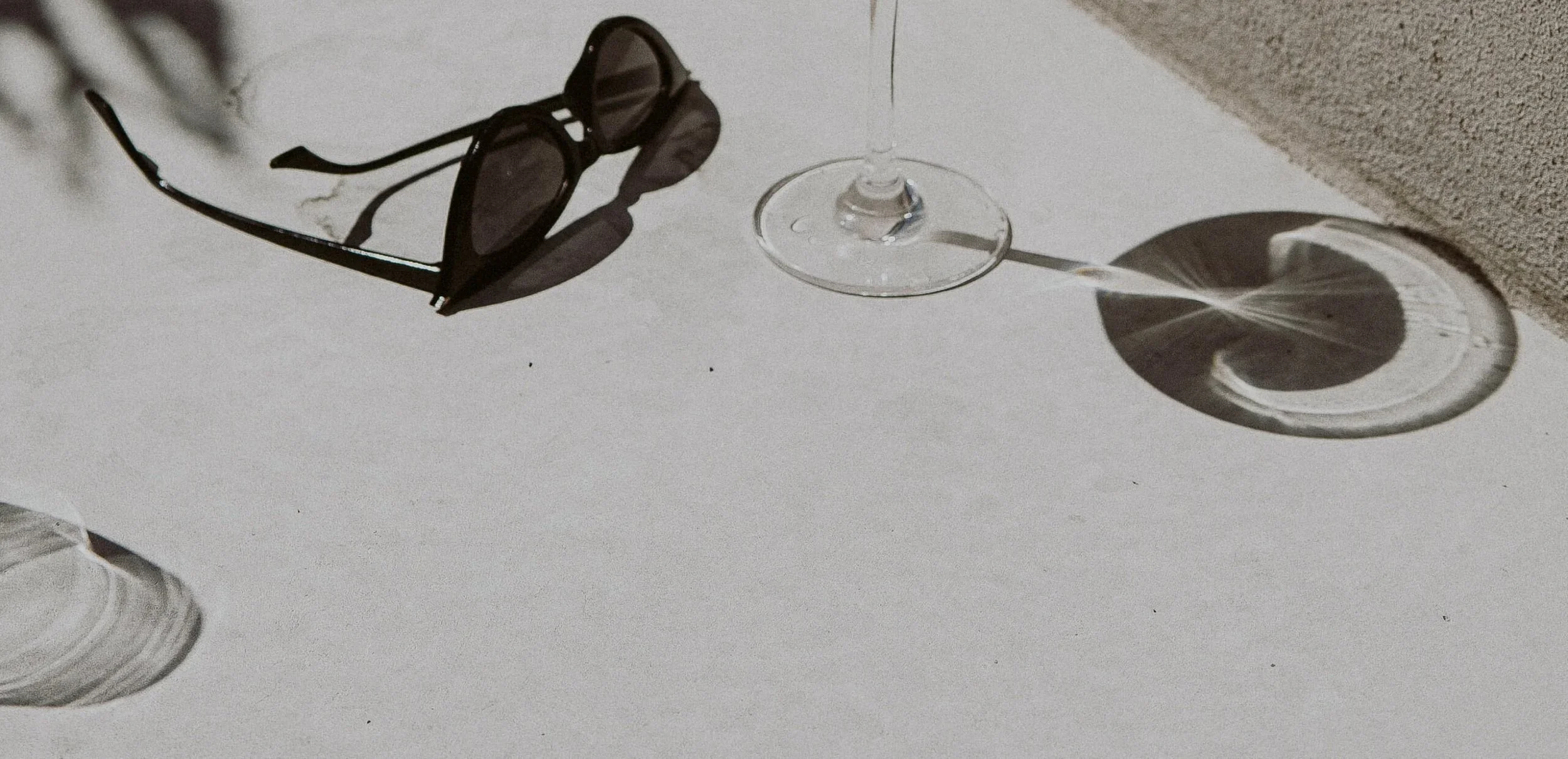 A pair of black sunglasses and an empty wine glass on a white surface, with shadows cast by the sunlight.