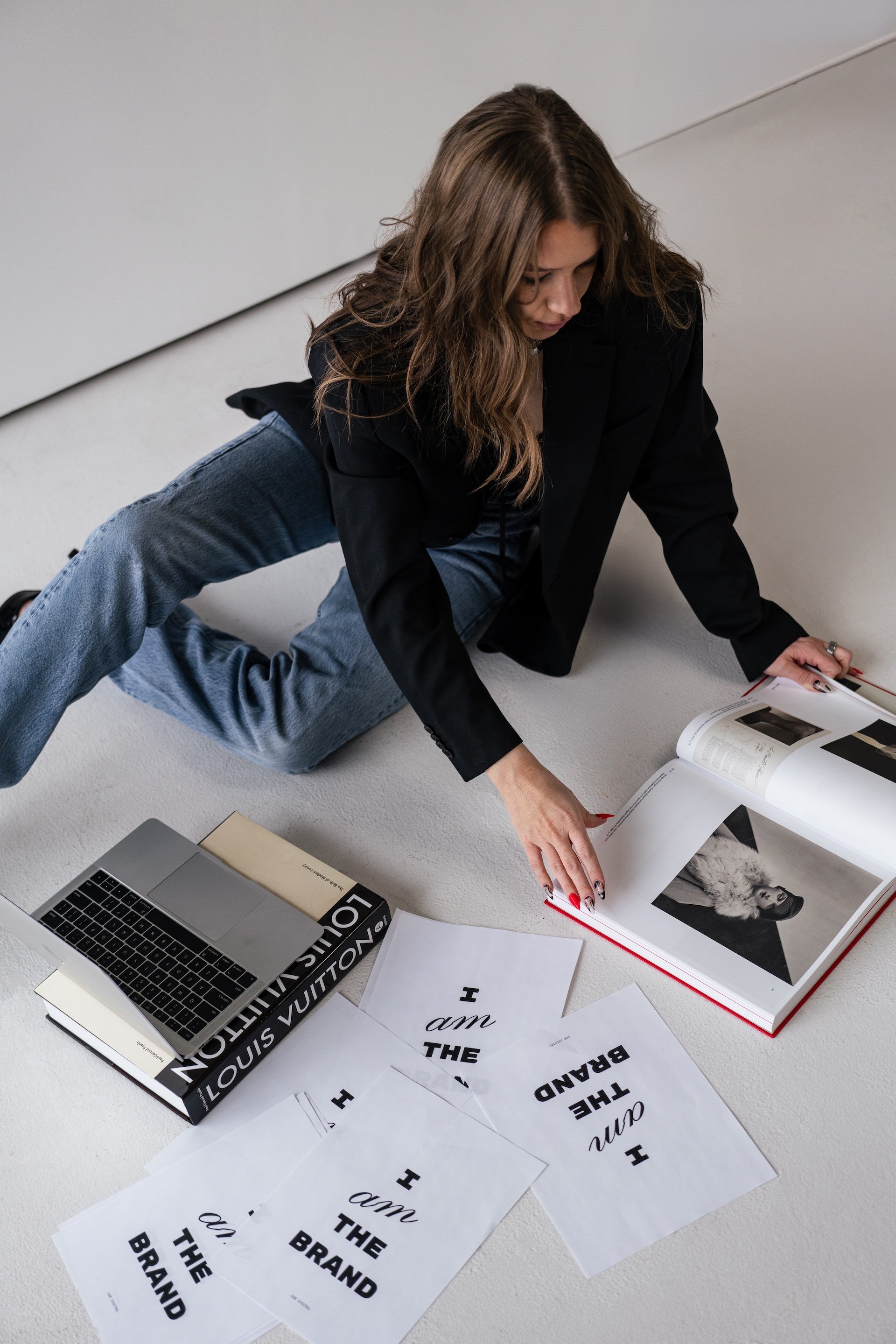 A woman with wavy brown hair is sitting on the floor, looking through a large photo book. She is wearing a black blazer and blue jeans. On the floor in front of her are printed sheets with phrases like 'I am the brand' and 'I am the way,' along with a silver laptop on a Louis Vuitton book.