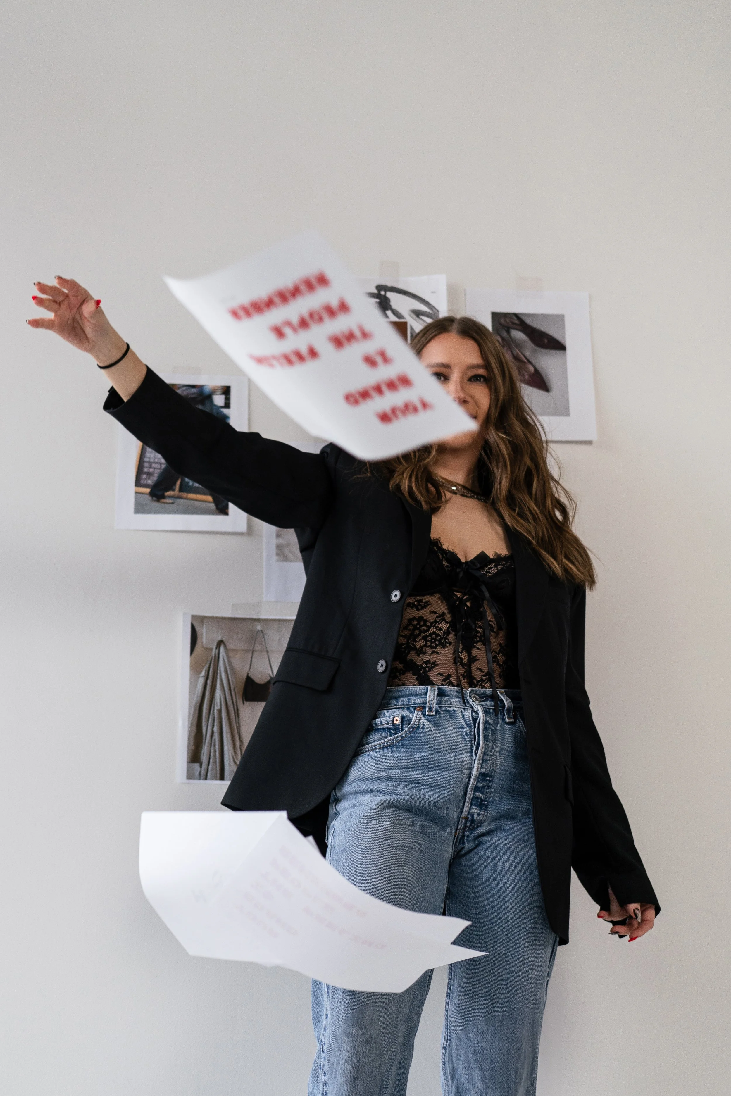 A young woman with brown hair in a black lace top and jeans, wearing a black blazer, tossing papers in the air in front of a white wall with fashion photos and illustrations.