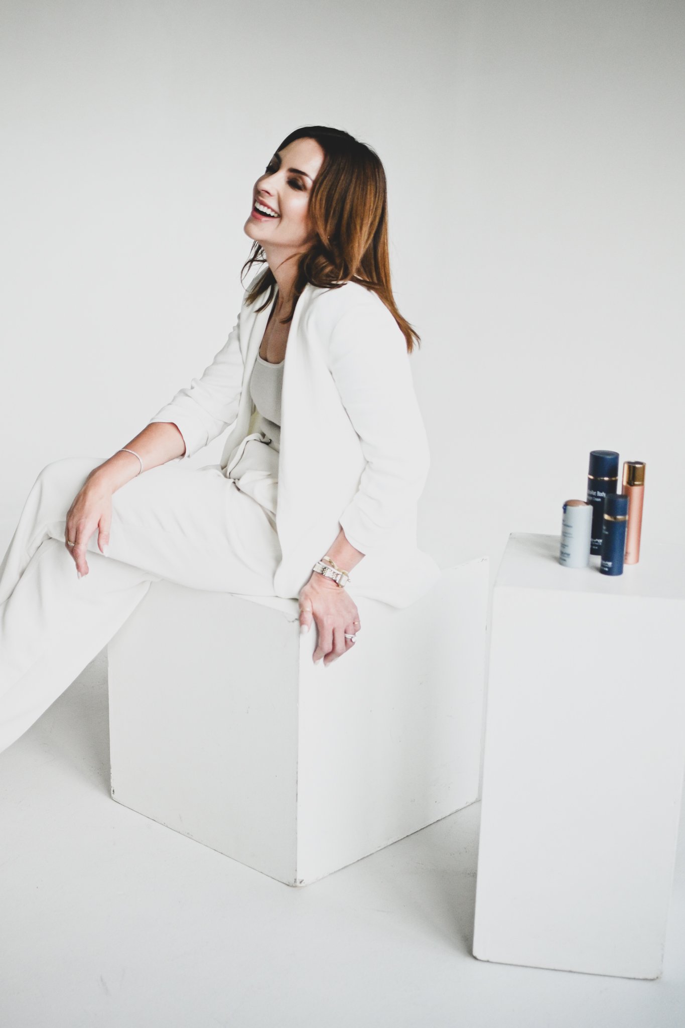 Woman in white sitting on a white block with a smile, beside a smaller white block with skincare products.