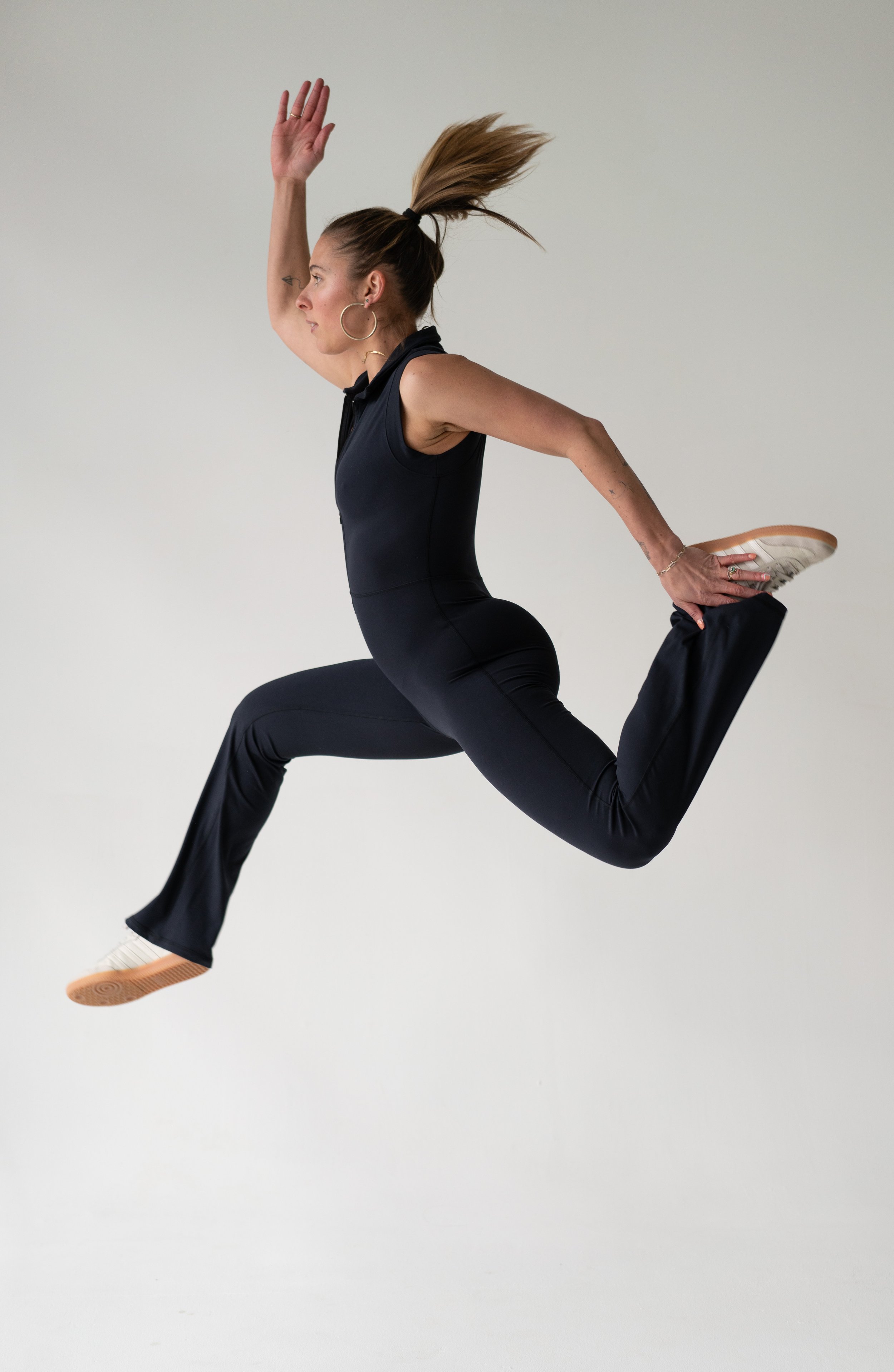 A woman jumping mid-air against a plain background, wearing black athletic clothing and sneakers.