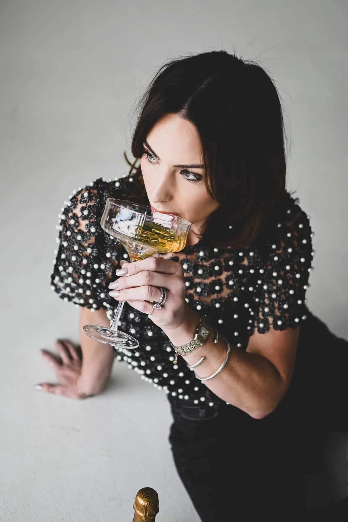 A woman in a black dress with pearl embellishments drinks champagne from a coupe glass.