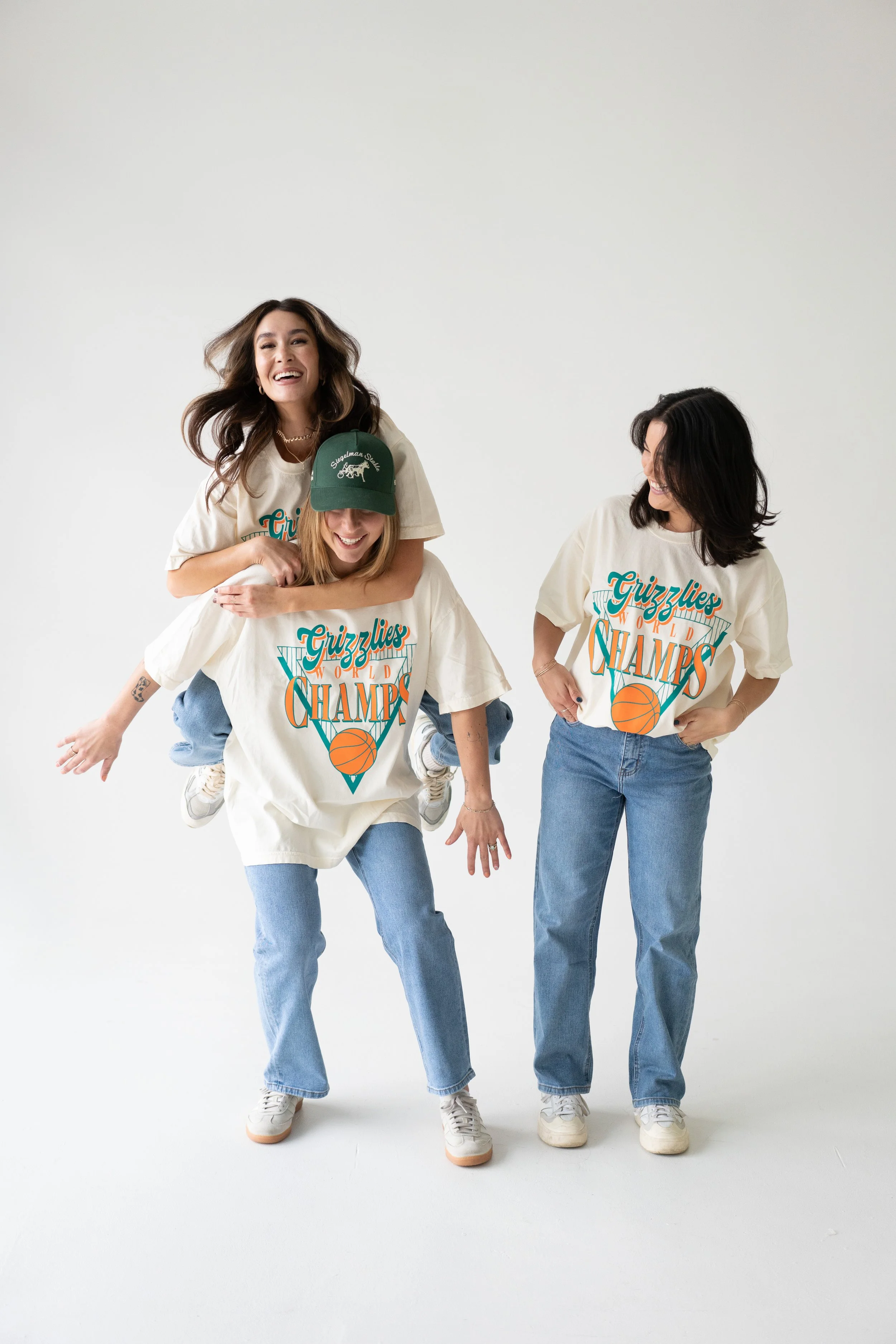 Three women smiling and having fun, with one giving a piggyback ride on the others. They are all wearing matching oversized t-shirts with a basketball graphic and the text 'Grizzlies World Champs', paired with jeans and sneakers, standing against a p
