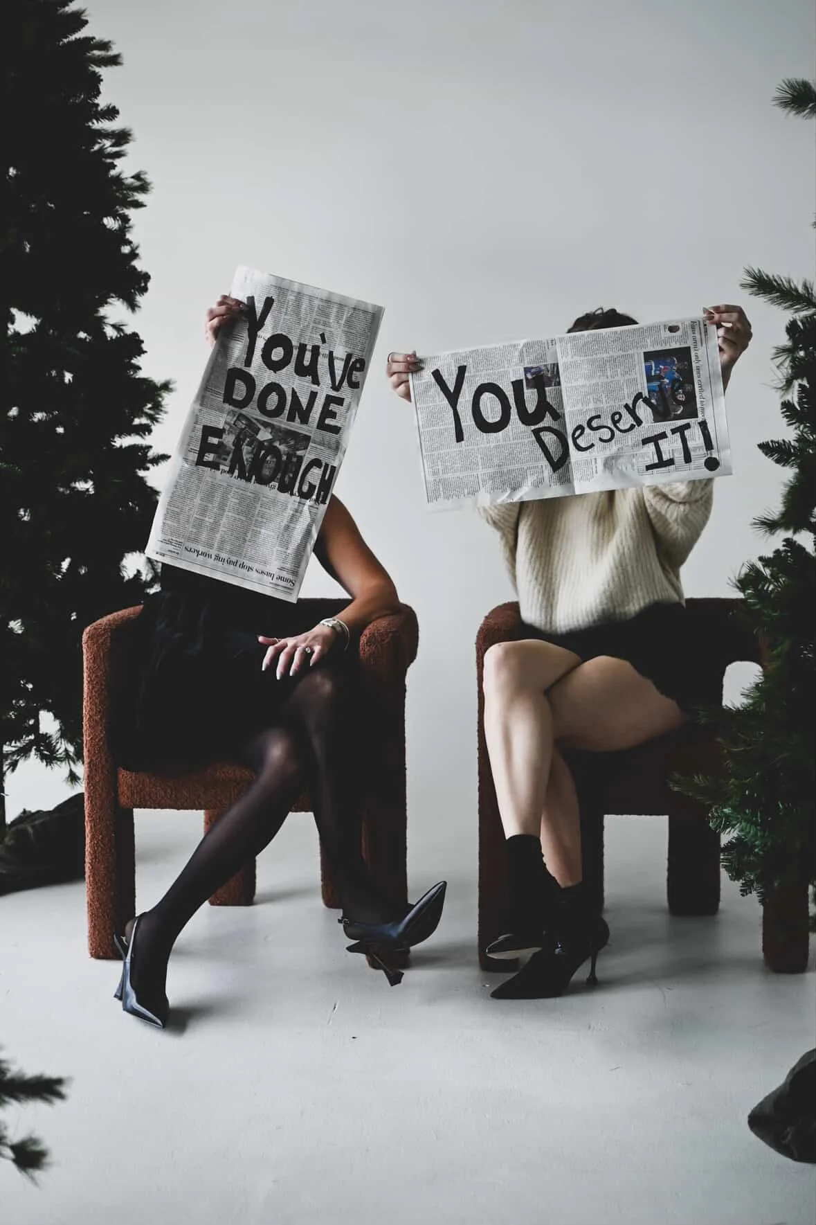 Two women sitting on chairs holding up newspapers with handwritten signs that say, 'You've Done Enough' and 'You Deserve It'. The women are dressed in dresses and heels, and there are Christmas trees partially visible in the foreground.