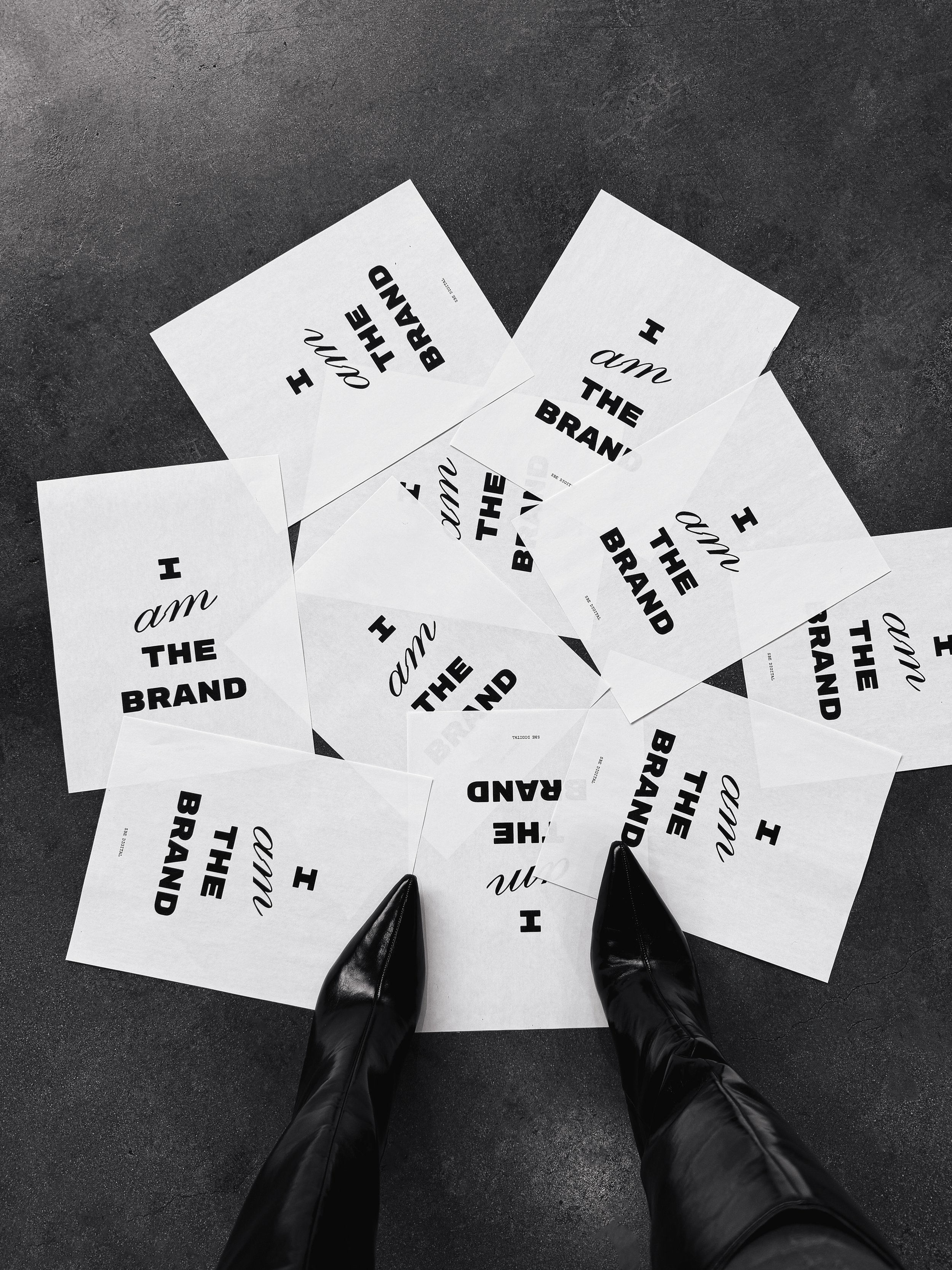 Multiple white sheets of paper scattered on a dark floor with the text "I am the brand" printed in bold black letters, viewed from above with black leather shoes visible at the bottom of the image.